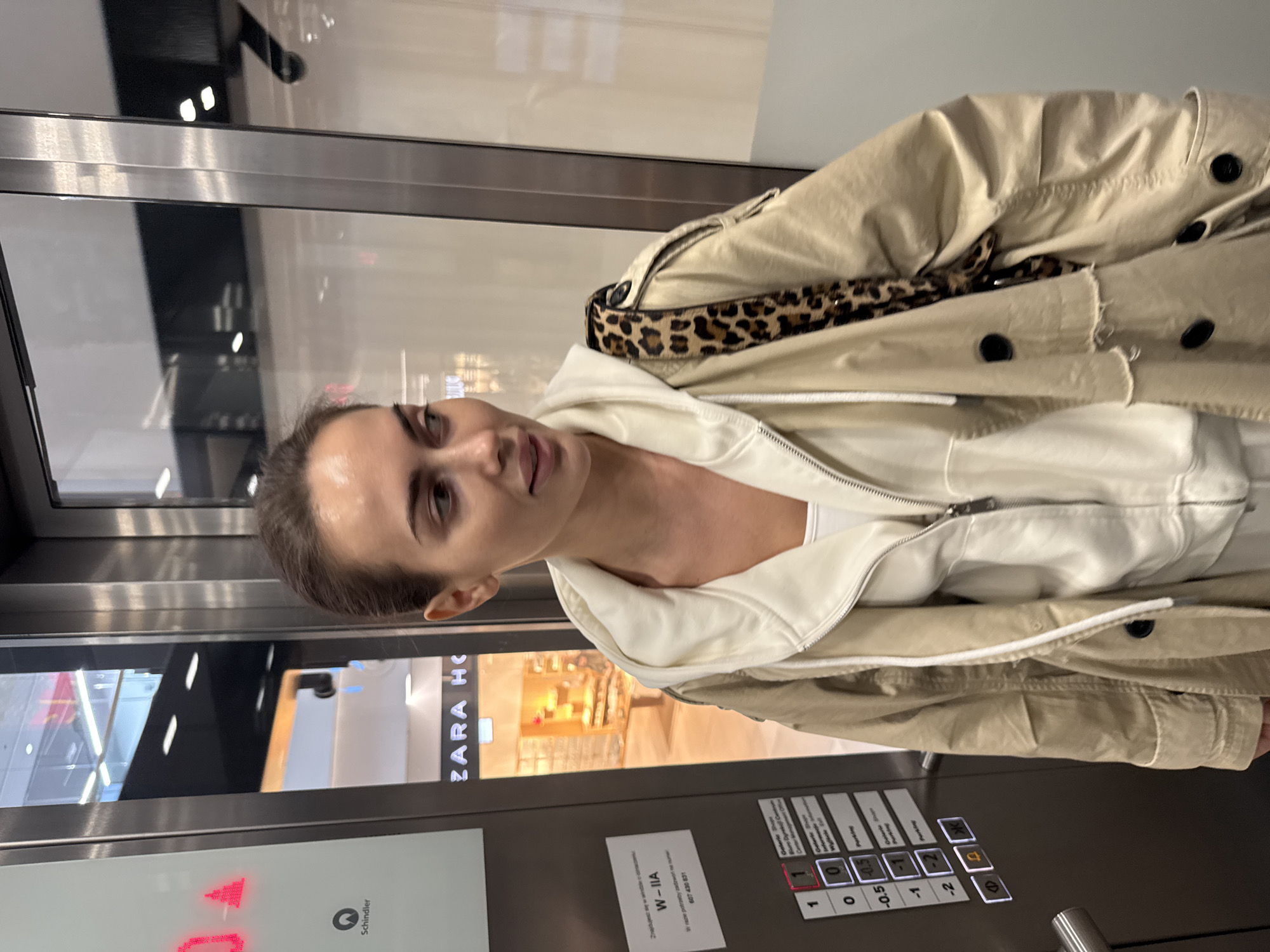 A young woman is depicted in a medium close-up, appearing to be in an elevator. She has dark hair pulled back, light eyes, and a natural, pleasant expression with a subtle smile. She is dressed in a light-colored, possibly cream or off-white, hooded sweatshirt, layered under a beige or khaki trench coat. A strap with a distinctive leopard print pattern is visible over her right shoulder.
The setting is clearly an indoor public space, likely a shopping mall in Warsaw, Poland, as indicated by the visible storefronts through the elevator's glass panels, including "ZARA HOME" and "ZARA HC". The activity taking place is a posed photograph or selfie within the elevator.
Notable details include the elevator's control panel on the right side, featuring illuminated buttons for various floors: 1, 0, -0.5, -1, and -2, along with text elements like "Parking" and "Galerie". A red arrow next to the "0" button indicates the current floor or direction. The "Schindler" logo, an elevator manufacturer, is also visible. Taped to the panel is a white notice with Polish text, "W - IIA" and "W razie potrzeby zadzwoń na numer 607 430 831" (translating to "In case of need call number 607 430 831"). The lighting suggests an indoor, commercial environment.