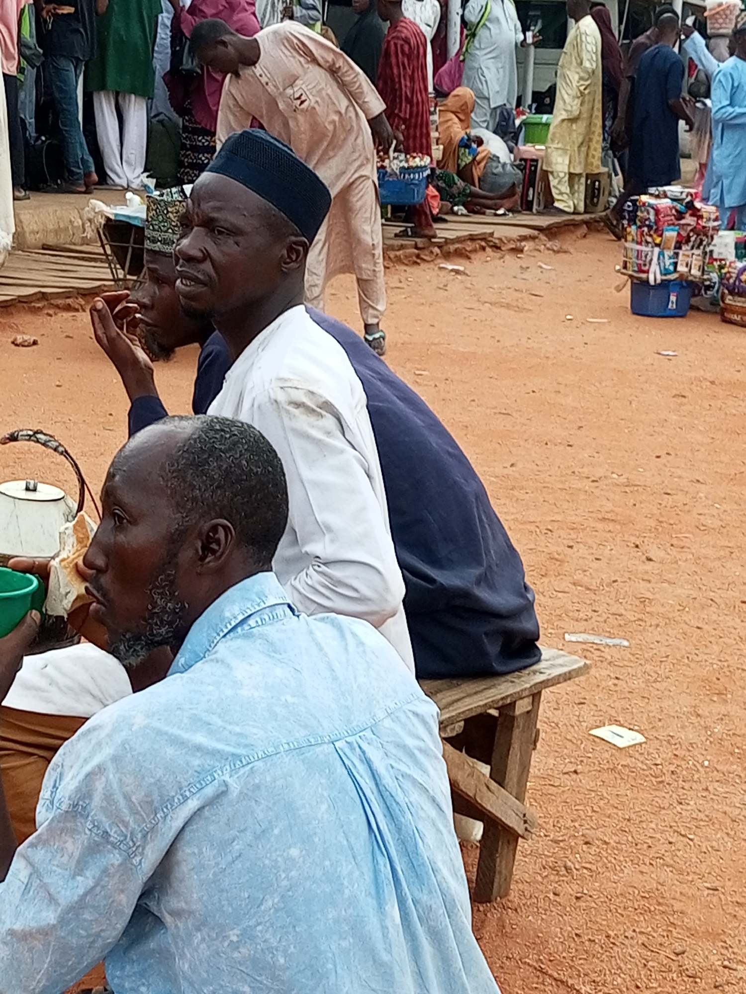 The image captures a street scene in the city of Gombe, Nigeria, likely in a market or gathering area. In the foreground, three men are visible. The man on the far left is wearing a light blue, short-sleeved collared shirt. He has a dark complexion and a greying, pointed beard. To his right, another man is partially visible, wearing a dark blue garment that covers his shoulder and back. The man in the center is the most prominent, wearing a dark blue skullcap and a white garment with dark blue trim. He is looking to his left with his mouth slightly open, and his right hand is raised near his face.
In the background, several other people are present, some in traditional attire such as flowing gowns and head coverings. There are market stalls or displays with various goods, including packaged items in blue baskets. The ground is covered with reddish-brown dirt. A wooden bench is situated in the mid-ground, with a part of it in the foreground. A white and metallic kettle is placed on a surface to the left of the central man. The overall atmosphere suggests a busy public space.