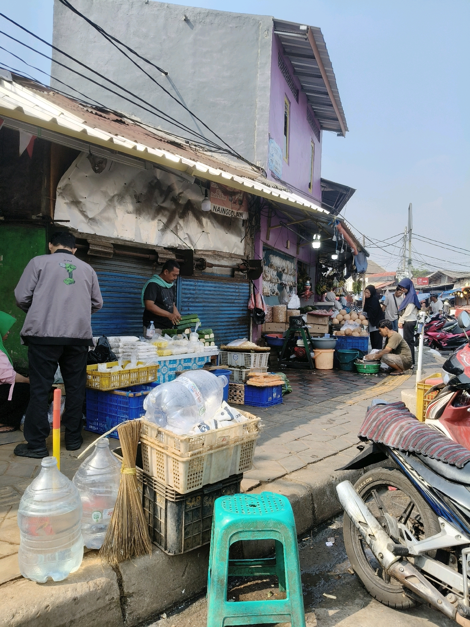 The image depicts a street scene in South Tangerang, Indonesia, likely a market area. In the foreground, two large plastic jugs of water are placed on a concrete curb, with a broom leaning against them. Next to the jugs are stacked crates containing various items, including what appears to be produce. A bright teal plastic stool sits nearby. Further back, a man in a grey jacket and black pants is standing, facing away from the camera, as if browsing or waiting. To his right, a vendor wearing a dark shirt and a light blue scarf is actively serving customers at a stall. The stall is laden with goods, including bundles of green leafy vegetables wrapped in plastic, and other packaged food items. Blue metal shutters partially cover the shop behind the stall. In the background, more market activity is visible, with people interacting, more stalls, and several motorcycles parked. A purple building with a light-colored roof stands out in the middle ground. The overall impression is that of a bustling outdoor market under a clear sky.