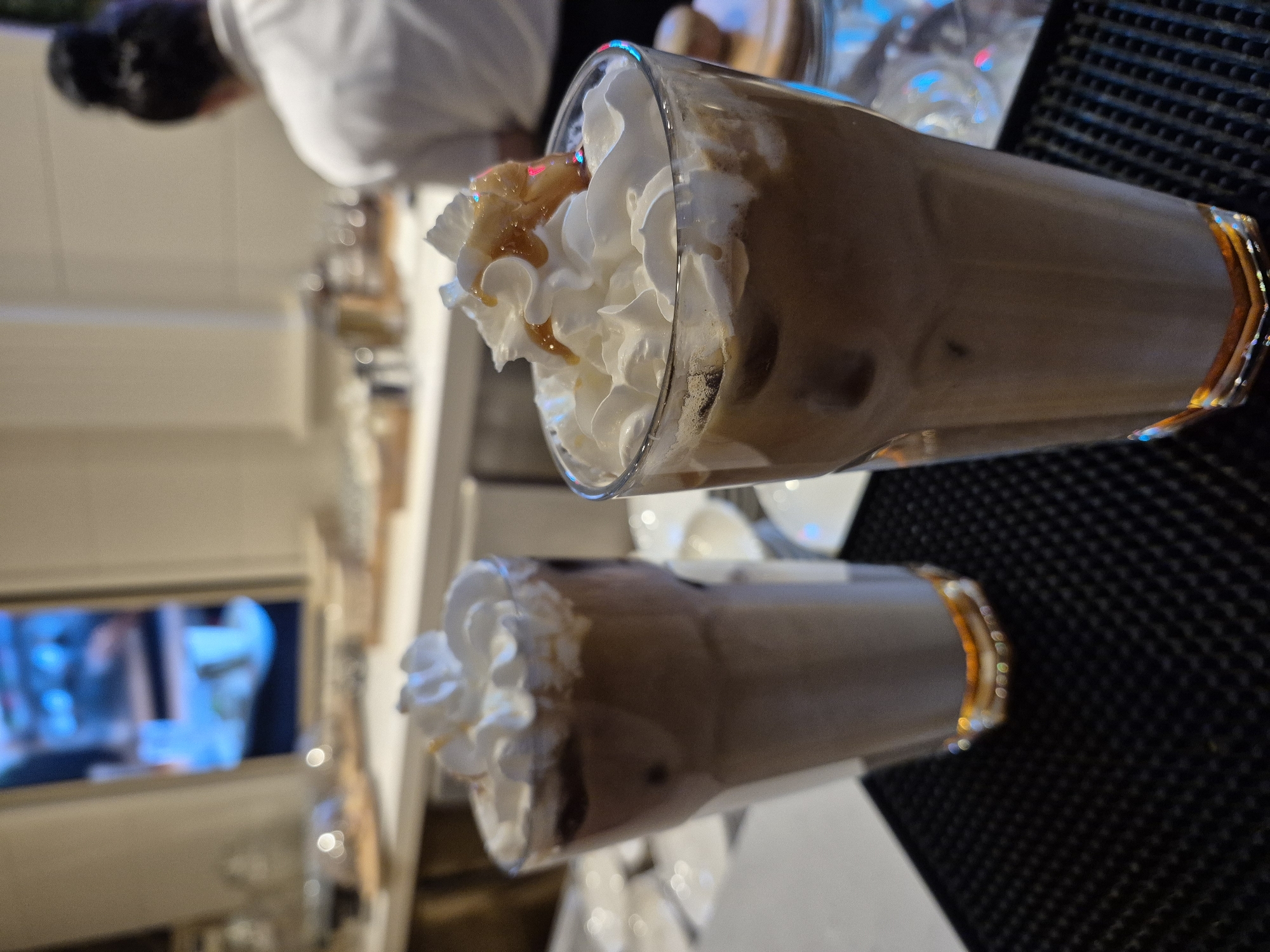 The image shows two tall glasses of iced coffee topped with whipped cream and caramel drizzle. The glasses are placed on a black surface with a textured pattern. In the background, a person with dark hair in a bun is partially visible, wearing a white shirt. A blurred shelf with various items and a TV screen displaying blue tones are also visible in the background. Based on the style of the cafe and drinks, as well as the mention of Alanya, Turkey, it can be inferred that the drinks are likely being served in a cafe or coffee shop in Alanya.