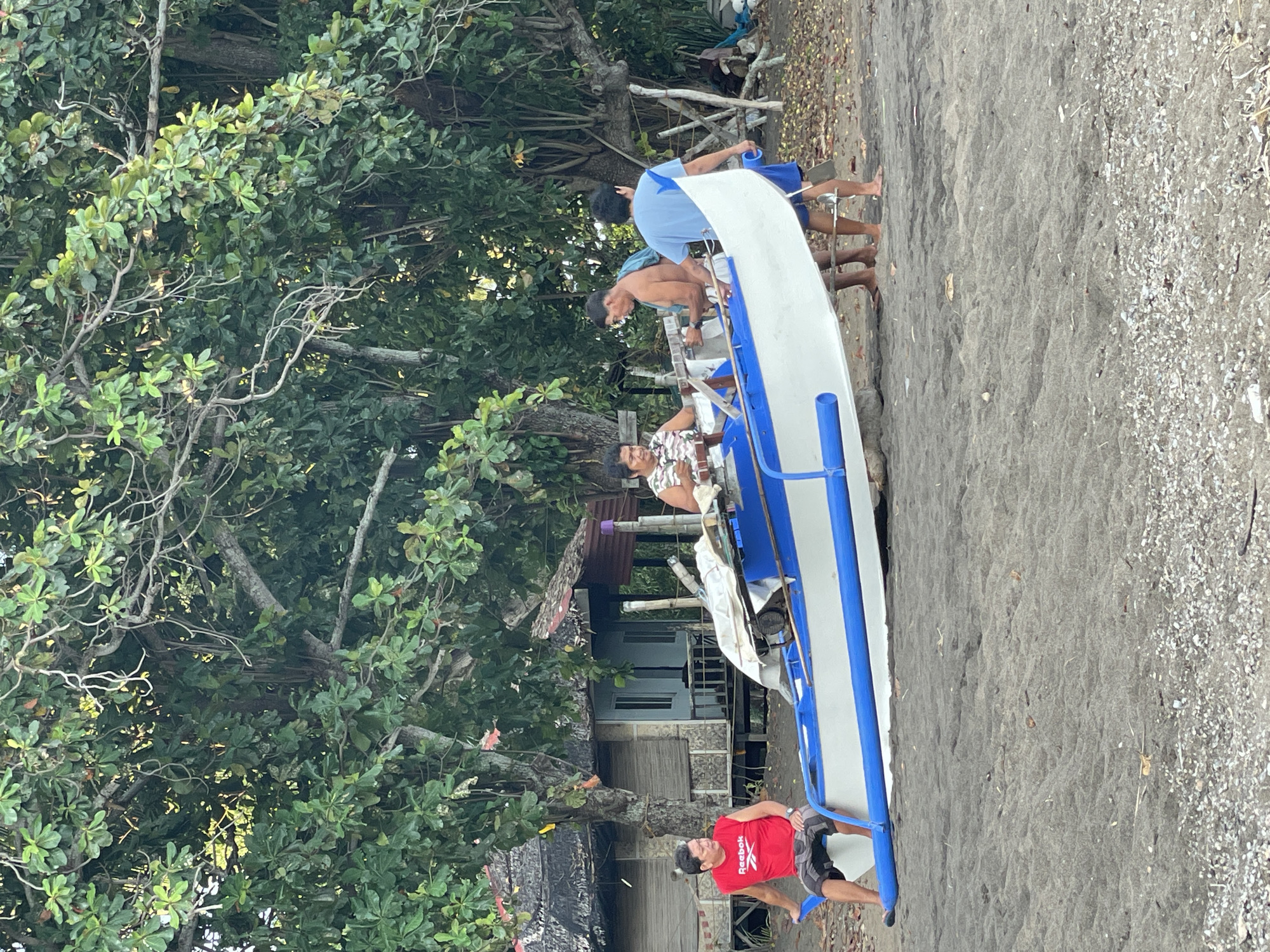 Four men are gathered around a white and blue outrigger canoe on a sandy beach in Dauin, Philippines. The canoe is positioned on the sand, angled towards the viewer's right.
The man on the far left, wearing a red t-shirt and patterned shorts, is seated on the edge of the canoe, facing left. He appears to be looking off to the side.
In the center of the image, a man with a patterned shirt is seated inside the canoe, facing left and holding what appears to be a musical instrument, possibly a ukulele. To his right, a bare-chested man is standing and looking at the canoe. Behind him, another man in a blue t-shirt and shorts stands, also looking towards the canoe.
Behind the men and the canoe, there is lush green foliage from large trees. In the background, to the left, a small, rustic structure with a thatched roof and shuttered windows is visible. The ground is covered in dark sand, with scattered small pebbles and debris. The overall scene suggests a relaxed, outdoor setting.