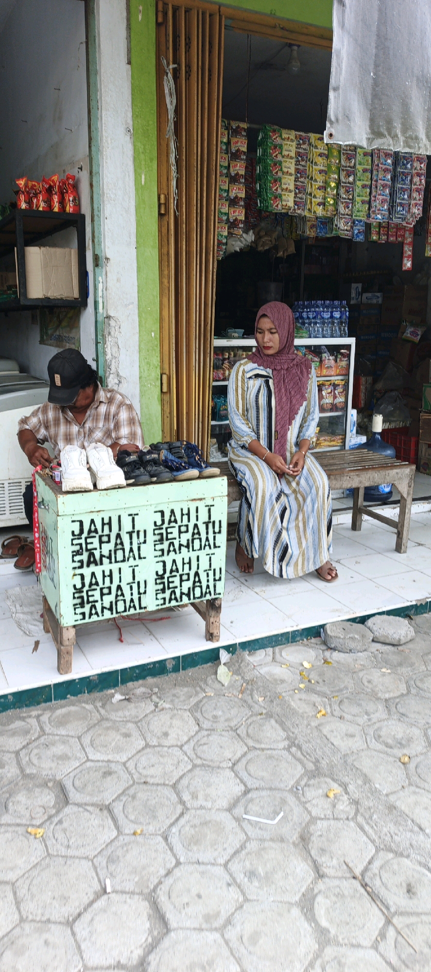 A man wearing a plaid shirt and a black baseball cap is seated on the left, focused on a light-colored shoe he is holding. In front of him, on a light green wooden stand, are several pairs of shoes and sandals. The stand has white lettering that appears to say "JAHIT SEPATU SANDAL" repeated. To the right of the stand, a woman wearing a maroon hijab and a striped dress is seated on a wooden bench, facing forward. She is looking downwards with her hands clasped. Behind them, a storefront is visible with a wooden accordion door. Shelves stocked with various packaged goods, including small sachets of food items and bottles, are displayed inside the store. The ground in front of the store is paved with hexagonal concrete tiles.