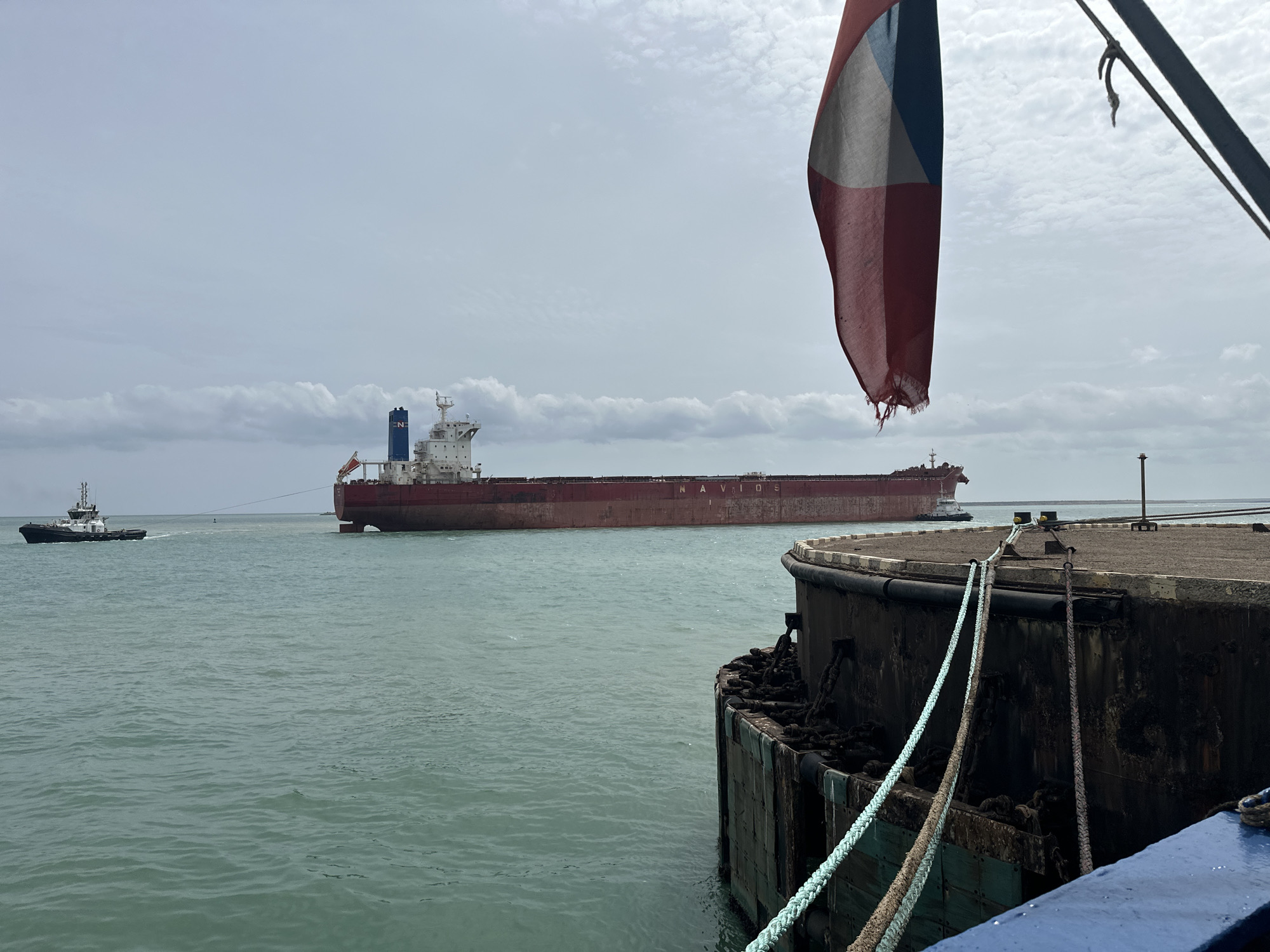 The scene captures a maritime operation under an overcast sky, viewed from a dock or another vessel. In the midground, a large, red-hulled bulk carrier, identified by the white letters "NAVIOS" on its side, is underway, moving from left to right. Its white superstructure, located towards the stern, includes a bridge, lifeboats, and a blue funnel bearing a white "N" emblem. The vessel appears lightly loaded, with a significant portion of its hull visible above the greenish-blue water. To the left of the bulk carrier, a smaller, dark-hulled tugboat with a white superstructure is actively pulling the larger vessel with a visible towline connected to its bow.
In the immediate foreground, to the right, a tattered, vertically draped tricolor flag displays stripes of red, white, and a dark blue or grey, with its bottom edge frayed. Below the flag and to the right, a heavily weathered concrete or metal dock structure is visible, featuring large chains draped over its side and prominent rubber fendering. Two mooring ropes, one light blue and one grey, are secured to this structure, extending out of the frame towards the viewer. A blue-painted edge of the structure from which the image is taken is also visible at the bottom right. The sky is uniformly overcast with light grey clouds. A faint vertical structure appears on the distant horizon to the far right. This scene is located in Uribia, Colombia.