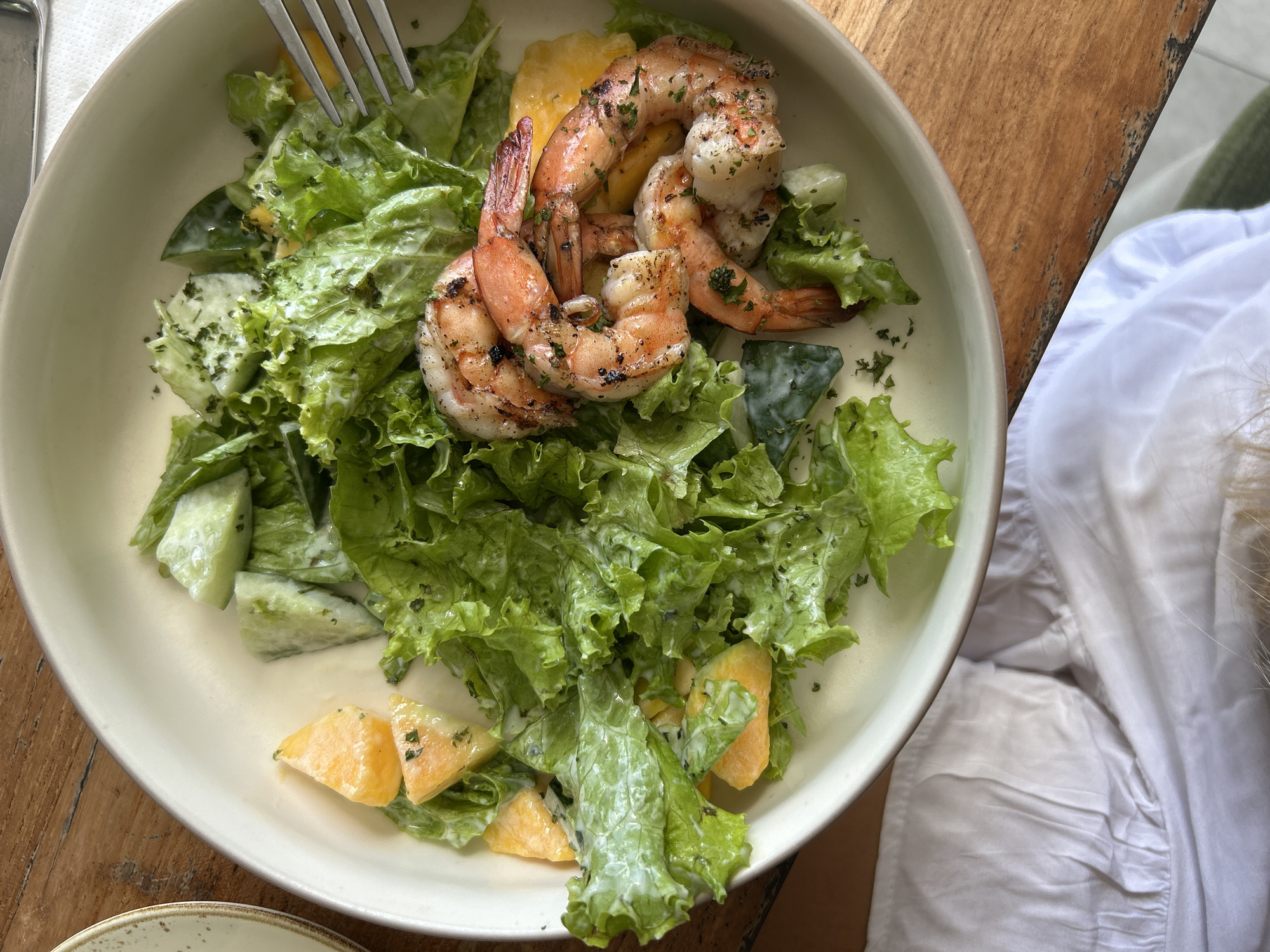 A bowl of salad is presented on a wooden table. The salad appears to consist of lettuce, shrimp, and sliced fruit, dressed with a creamy sauce. A fork is piercing the salad. A white cloth and a glimpse of a person's hair are visible to the right of the bowl. The image was likely taken in Kuta Selatan, Indonesia.