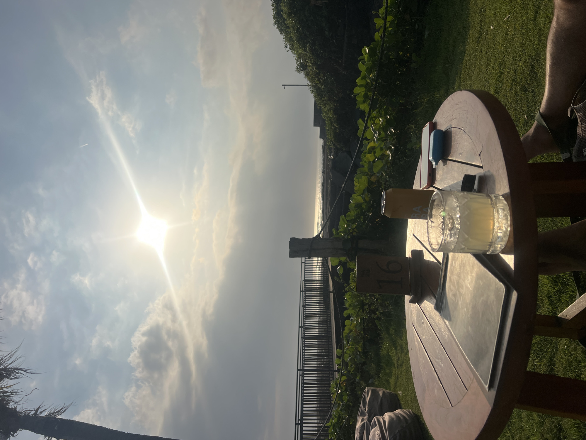 In Mengwi, Indonesia, at an outdoor restaurant with a view of the ocean, a round wooden table is set. On the table is a glass of a light-colored beverage with ice, a can of beer, a tablet, a mobile phone, and a small blue electronic device. A wooden table number stands prominently, displaying the number "16." The table is surrounded by lush green grass. In the background, a boardwalk leads to the beach, with the ocean stretching to the horizon under a partly cloudy sky illuminated by the setting sun. Beanbag chairs are visible to the left of the table, and dense green foliage lines the area near the boardwalk. A person's legs are visible on the right side of the frame, indicating their presence at the table.