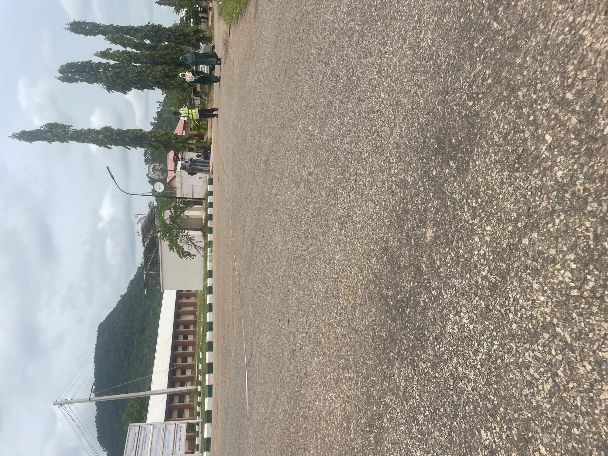 The image depicts a view of a road in Lokoja, Nigeria, with a mountain range in the background. In the foreground, the asphalt road fills the majority of the frame. To the left, a white building with a brown roof and columns is visible, with a signpost in front of it. Behind this building, a steep, tree-covered mountain rises against a cloudy sky. Further to the right, several tall, slender trees stand prominently. In the middle ground, a group of people are gathered. One individual, wearing a high-visibility vest, stands near the center. To their right, two women in hijabs are walking. Other individuals are visible in the background near buildings. The environment appears to be an outdoor, possibly institutional or commercial, setting, with elements of natural landscape and urban infrastructure.