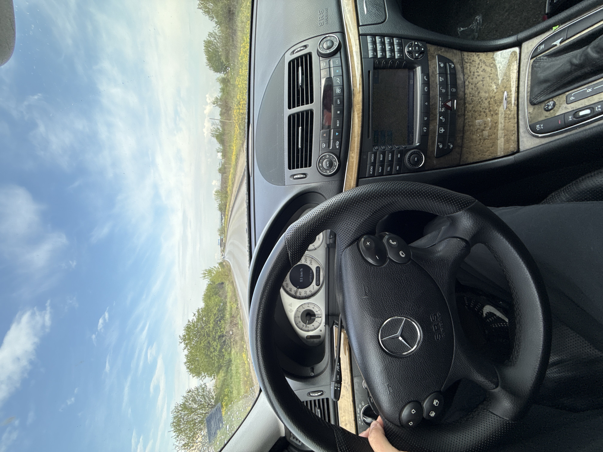 The image shows the interior of a Mercedes-Benz car. The dashboard is visible, along with the steering wheel, instrument panel, and center console. The exterior is visible through the windshield, showing a road with greenery. The speedometer indicates a speed of 53 km/h. The location is unknown and cannot be determined from the given information.