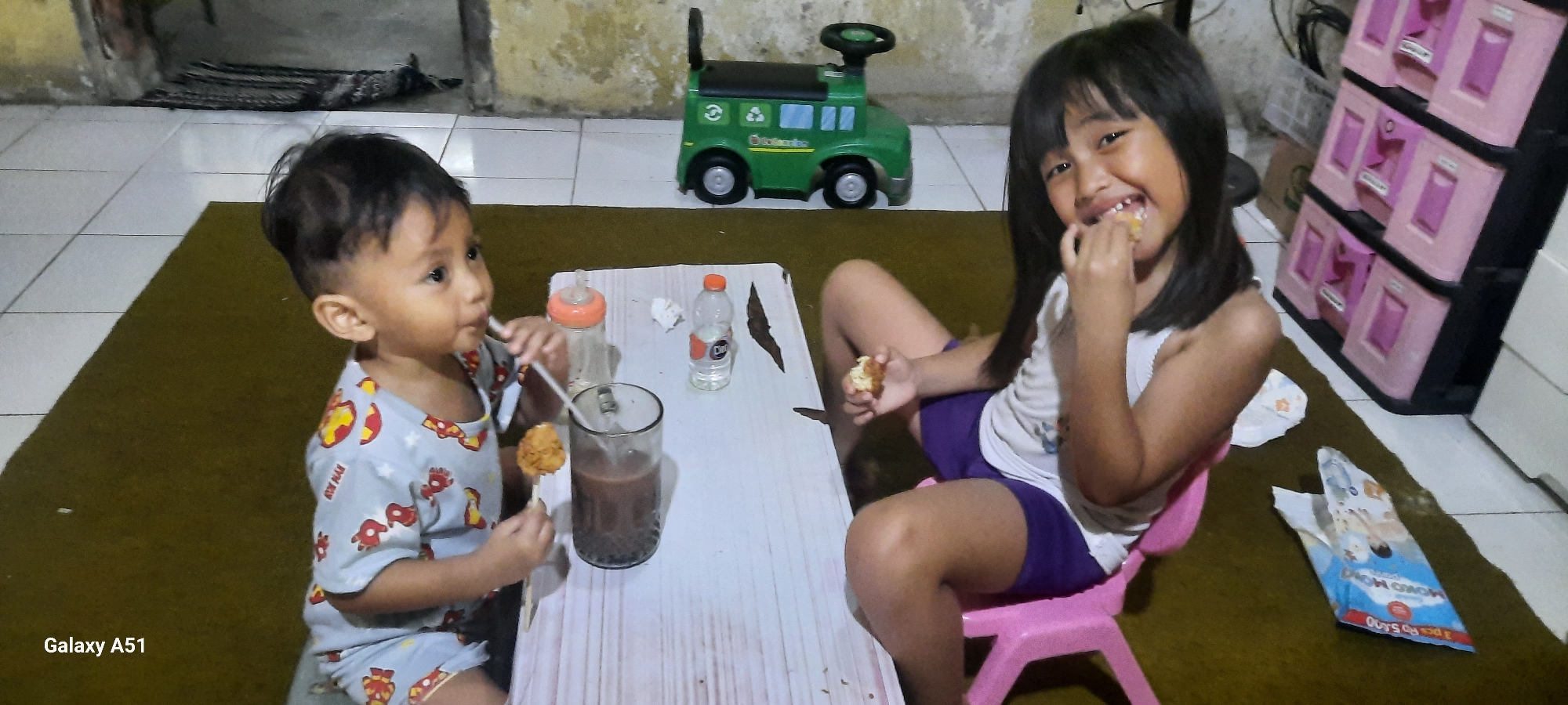 In the city of Jatitengah, Indonesia, a young boy and girl are seated at a low white table on a brown mat. The boy, wearing a light blue shirt with red and yellow cartoon prints, holds a stick with a round brown food item and a straw in a glass of chocolate-colored liquid. The girl, wearing a white top and purple shorts, is eating and smiling. On the table, a baby bottle with an orange nipple and a plastic water bottle are visible. Behind the children, a green toy vehicle with recycling symbols is present. To the right of the scene, a pink child-sized chair and a stack of pink plastic drawers are in the background, with a crumpled blue and white package on the floor. The environment appears to be indoors, with tiled flooring and a textured wall.