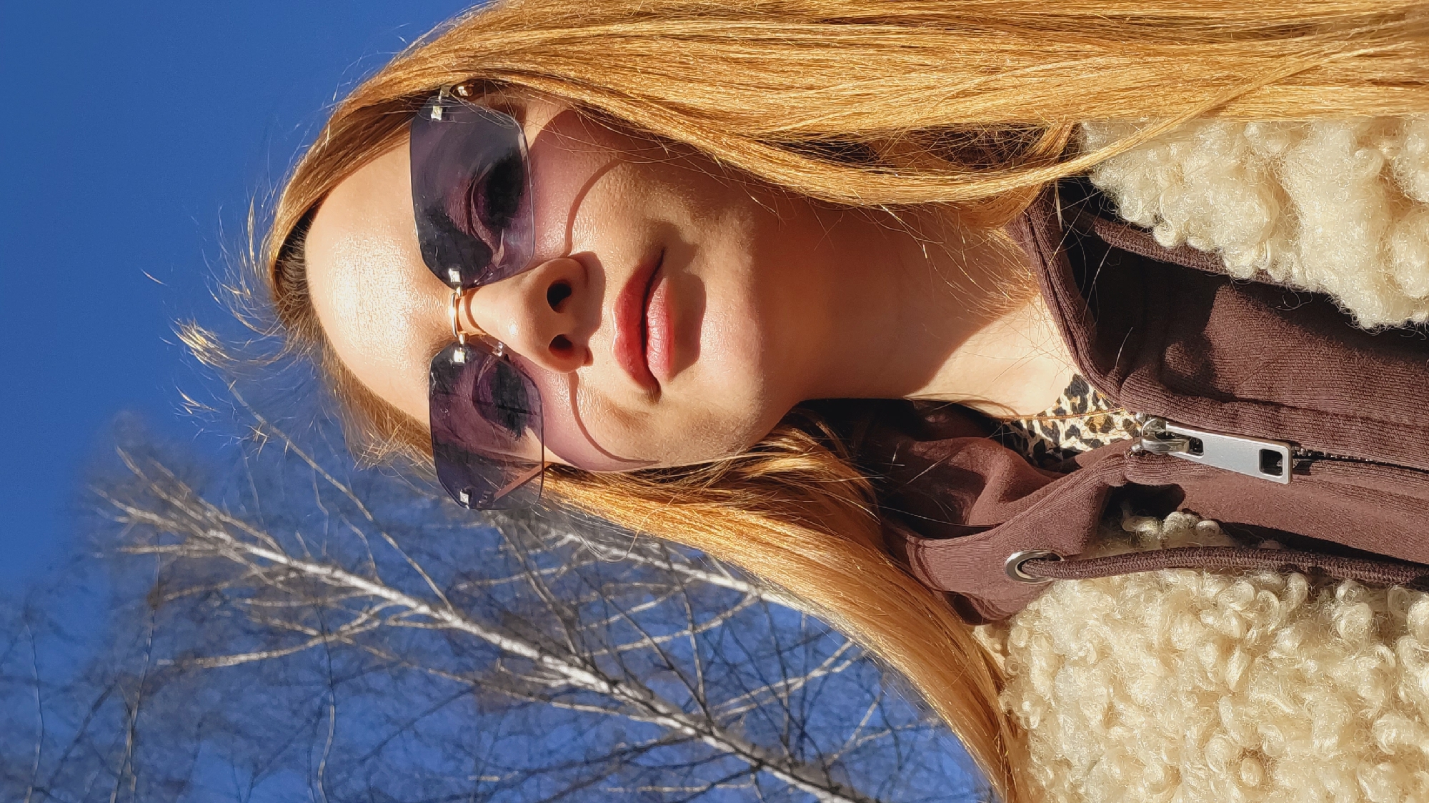 A young woman with long, straight, reddish-blonde hair is wearing sunglasses in an outdoor setting. Her face is partially illuminated by sunlight, casting shadows across her nose and cheek. She is wearing a brown hooded jacket with a cream-colored sherpa lining. The jacket is partially zipped up, revealing a leopard-print top underneath. The background shows a bright blue sky and the bare branches of trees, suggesting a location in or near Chaikovsky, Russia, possibly during late fall or early spring. The photo is taken from a low angle, tilting upwards towards the subject.