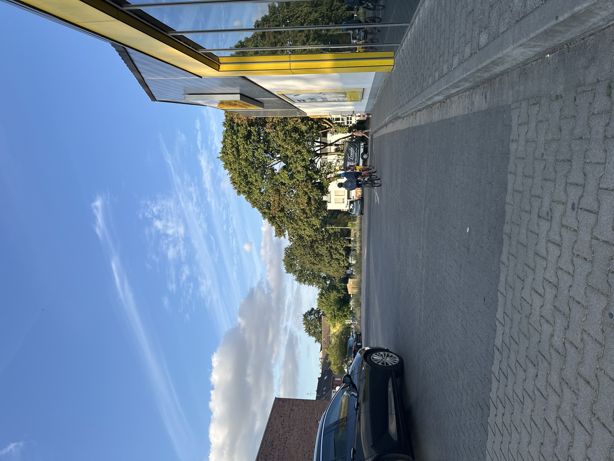 This image depicts an outdoor scene in the city of Krefeld, Germany, likely a parking lot adjacent to a commercial building. A dark-colored station wagon is parked on the left side of the frame, its rear half visible. In the mid-ground, a group of people on bicycles are moving away from the camera. The building on the right features a yellow and grey facade with large reflective windows. A sidewalk or paved area separates the parking lot from the building. Trees and other buildings are visible in the background under a partly cloudy blue sky.