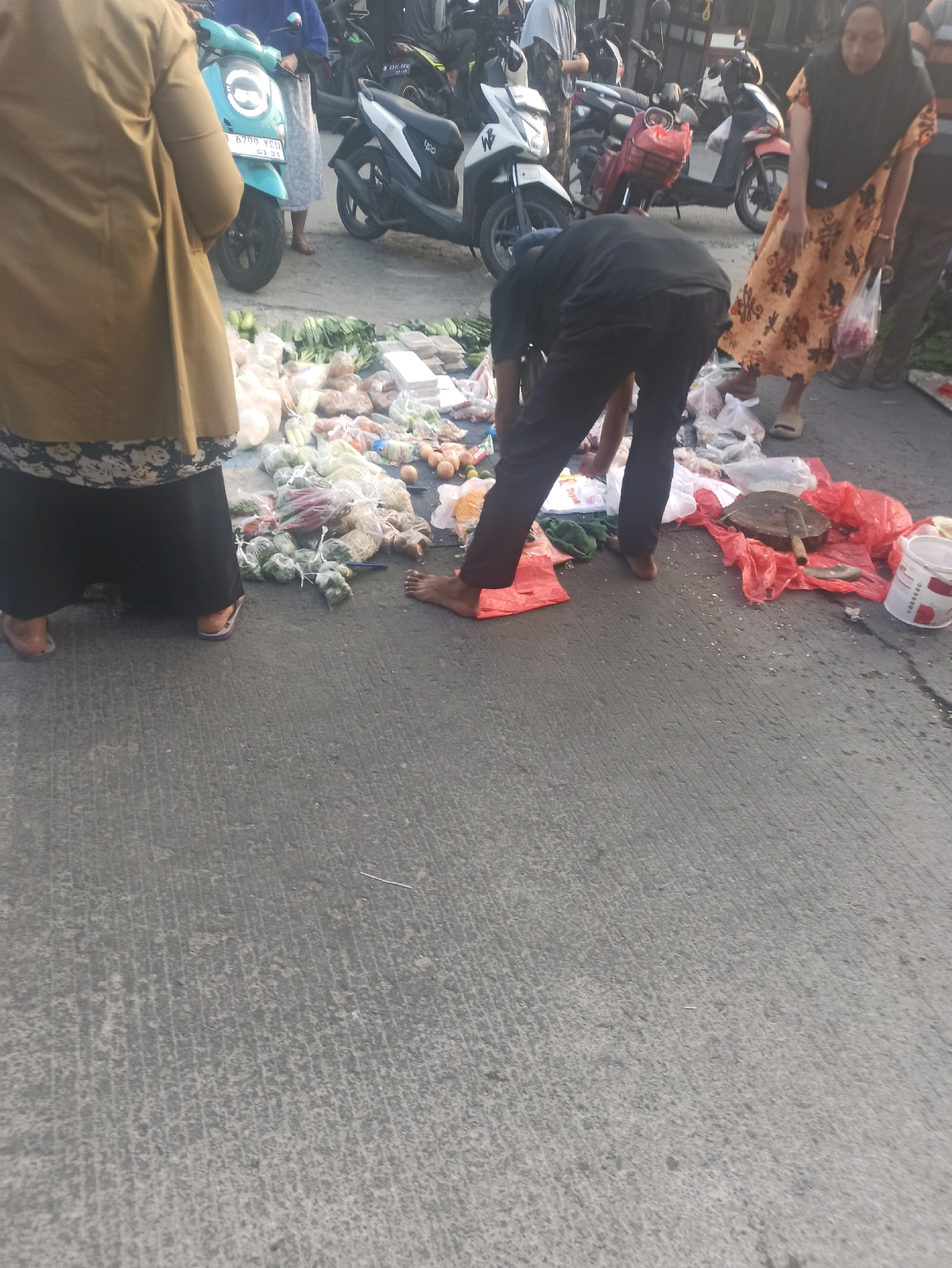 In Telagamurni, Indonesia, a street market scene unfolds. The foreground shows a person in dark clothing bending over, sorting through produce laid out on a dark tarp on the asphalt. They are barefoot and wearing dark pants. Scattered around them are various fruits and vegetables in plastic bags, along with some loose items like eggs. To the left, a person in a tan jacket and patterned skirt stands, facing away from the camera. Behind them, a turquoise scooter is parked. In the background, several other motorcycles, including a white and black model and a red one, are visible. A woman in a floral dress and hijab stands to the right, holding a plastic bag. The ground is asphalt, marked with some debris and a white bucket. The overall environment appears to be an outdoor market with a somewhat cluttered arrangement of goods and vehicles.