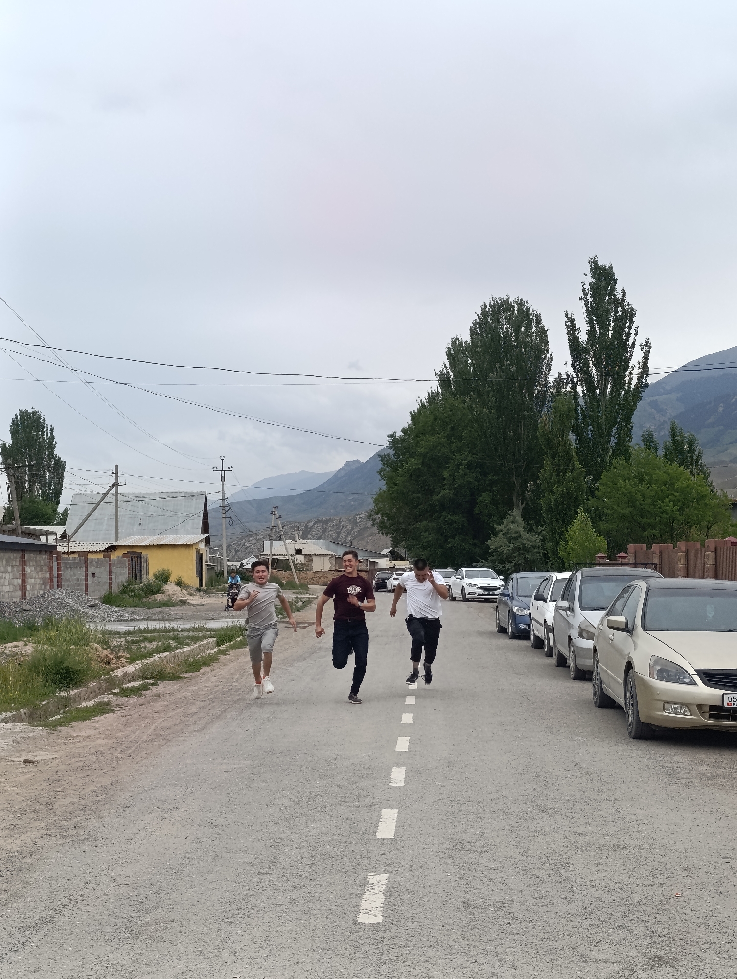 In Naryn City, Kyrgyzstan, three young males are captured in mid-stride as they run down a paved road. The person on the left wears a light gray t-shirt and matching shorts with white sneakers. The middle runner is dressed in a maroon t-shirt and dark pants with white accents on his shoes. The third runner, on the right, sports a white t-shirt and black pants with black sneakers. They are running away from the camera on a road that appears to be a two-lane street with a dashed white line marking the center. On the right side of the road, a line of parked cars is visible, including a light-colored sedan in the foreground displaying a license plate. The left side of the road features a low brick wall, some construction materials, and overgrown vegetation. In the background, there are buildings, tall trees, power lines, and mountains under a cloudy sky. The overall scene depicts an outdoor activity in a semi-urban or rural setting.