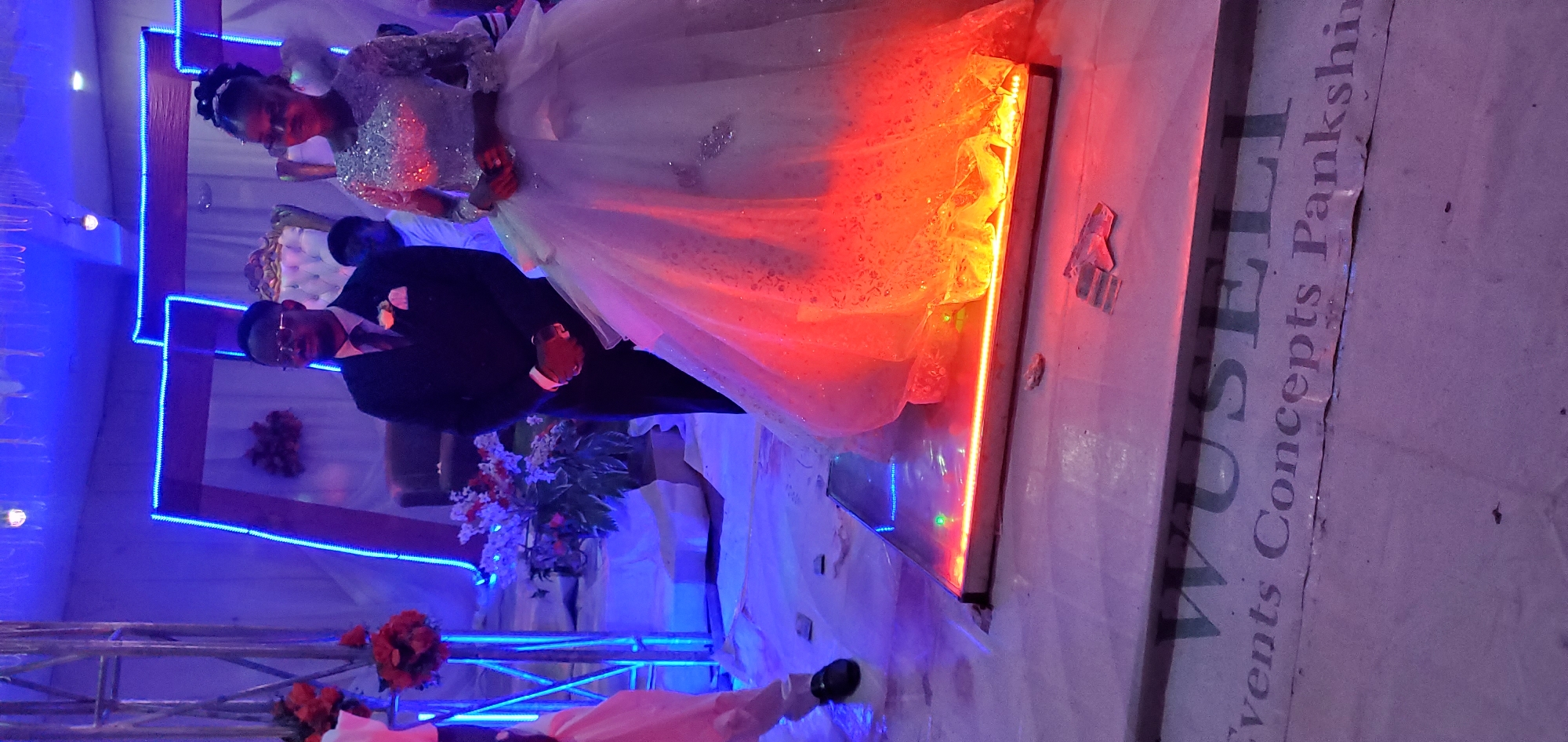This image captures an indoor event, likely a wedding reception, in Lankan, Nigeria. Two central figures, identified as a bride and groom, stand formally on an illuminated rectangular stage platform.
The groom, positioned on the left, is a male wearing a dark suit over a light-colored shirt and a dark tie. A light-colored boutonnière is affixed to his left lapel. He wears glasses and stands with his hands clasped in front of him.
The bride, positioned on the right, is a female wearing a shimmering, light-colored gown with a full, voluminous skirt and long, embellished sleeves. The lower portion of her gown is vibrantly illuminated by a strong red-orange light emanating upwards from within the stage platform. Her hands are also clasped in front of her.
A third male individual is partially visible behind the couple, wearing a light-colored top. To the far left, the lower leg and foot of another person, wearing a dark shoe and light-colored trousers, are visible on the floor adjacent to the stage.
The venue is adorned with celebratory decorations and lighting. Two large, structural frames, possibly arches, are prominent in the background, outlined with bright blue LED strips. A white draped backdrop is visible behind the frames, and ornate, light-colored chairs are partially discernible. Floral arrangements, including a bouquet of red flowers on a stand to the left and a plant with white and red flowers near the stage, enhance the decor.
The floor surrounding the stage is covered with a light-colored material. Scattered small, rectangular pieces of material are visible on the floor, particularly near the stage. Text printed on the floor covering at the bottom of the image reads "WUSELI Events Concepts Pankshin".
The lighting is predominantly artificial, creating a dynamic ambiance with blue ambient light from overhead and background elements, contrasted sharply by the red-orange glow originating from the stage platform that highlights the bride's gown. A single bare light bulb is visible hanging from the ceiling.
