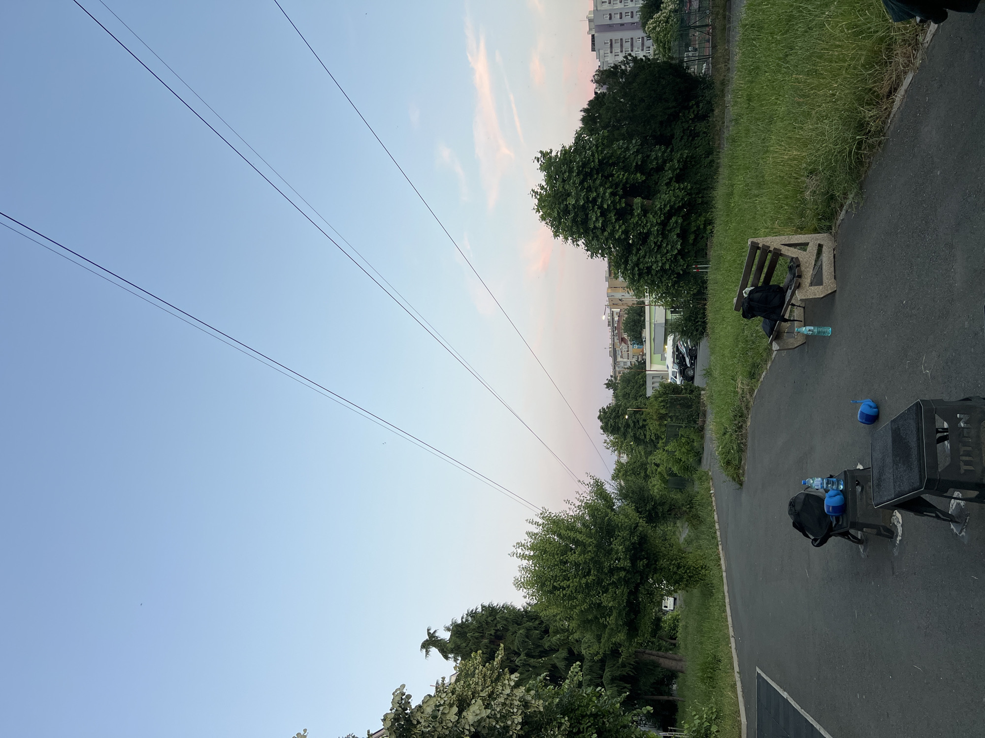 The image depicts an outdoor park or recreational area in Burgas, Bulgaria, as evidenced by the presence of power lines, residential buildings, and the specific flora. The scene is set during twilight, with a clear sky transitioning from light blue to a soft pink hue. Three prominent power lines stretch across the upper portion of the frame, angled from the left to the right.
In the foreground, a paved pathway runs through the park. On the pathway, several items suggest recent or intended physical activity. Two black, sturdy boxes, possibly exercise equipment or benches, are placed on the pavement. One box is closer to the viewer, while the other is further back. On the further box, a black backpack and a blue water bottle are visible. To the right of these boxes, a wooden bench with concrete supports is situated. A black backpack and a blue water bottle are also placed on this bench. On the ground near the boxes, a pair of blue weighted gloves are lying.
Beyond the pathway, a strip of vibrant green grass extends, lush and somewhat overgrown in places. Behind the grass, a dense cluster of trees and foliage forms a natural barrier. In the mid-ground, more trees are visible, with a road and parked cars suggesting proximity to a residential or urban area. Residential buildings, characterized by multi-story structures, are visible in the background, with some displaying light-colored facades and others with grey and purple accents. The overall atmosphere is one of quietude, with no people visible in the scene.