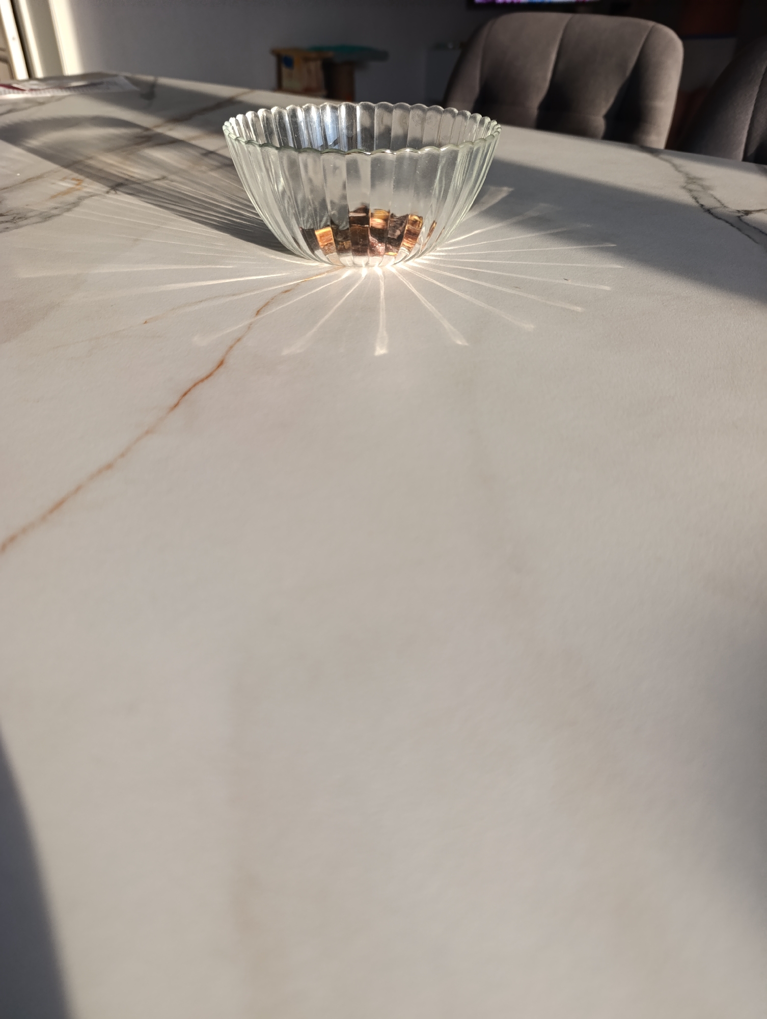 A glass bowl sits on a marble-like table, casting intricate shadows from the sunlight. The bowl appears to be filled with small, irregularly shaped pieces, possibly candies or decorative stones, which are catching the light. The table's surface has a subtle veining pattern, with a reddish-brown streak running across it. In the background, out of focus, are two upholstered chairs with a tufted design, suggesting a dining or living area. A hint of furniture, possibly a stool or small table, is visible further in the background, along with what looks like blinds or shutters on a window, from which the light is streaming. The lighting indicates it is daytime, likely late afternoon given the strong, angled sunlight creating the dramatic shadows. The scene is indoors, in a well-lit room. No people are visible in the frame.