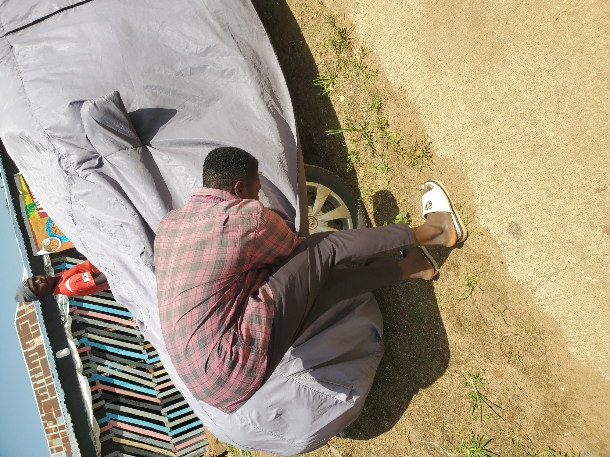 In an outdoor environment in Unknown, Nigeria, a person wearing a red and black checkered shirt and dark pants is bent over, partially obscured by a large, light-colored car cover. The person's head is down, and they appear to be working on or near the front wheel of a vehicle underneath the cover. Another person, wearing a grey beanie and an orange shirt, stands to the left of the frame, looking towards the viewer. In the background, there is a structure with a corrugated metal roof and a colorful wooden fence or partition made of vertically aligned planks in shades of pink, blue, and white. The ground is a mixture of dirt and some sparse green grass, with a concrete surface visible in the lower right of the image. The sky is clear and blue.