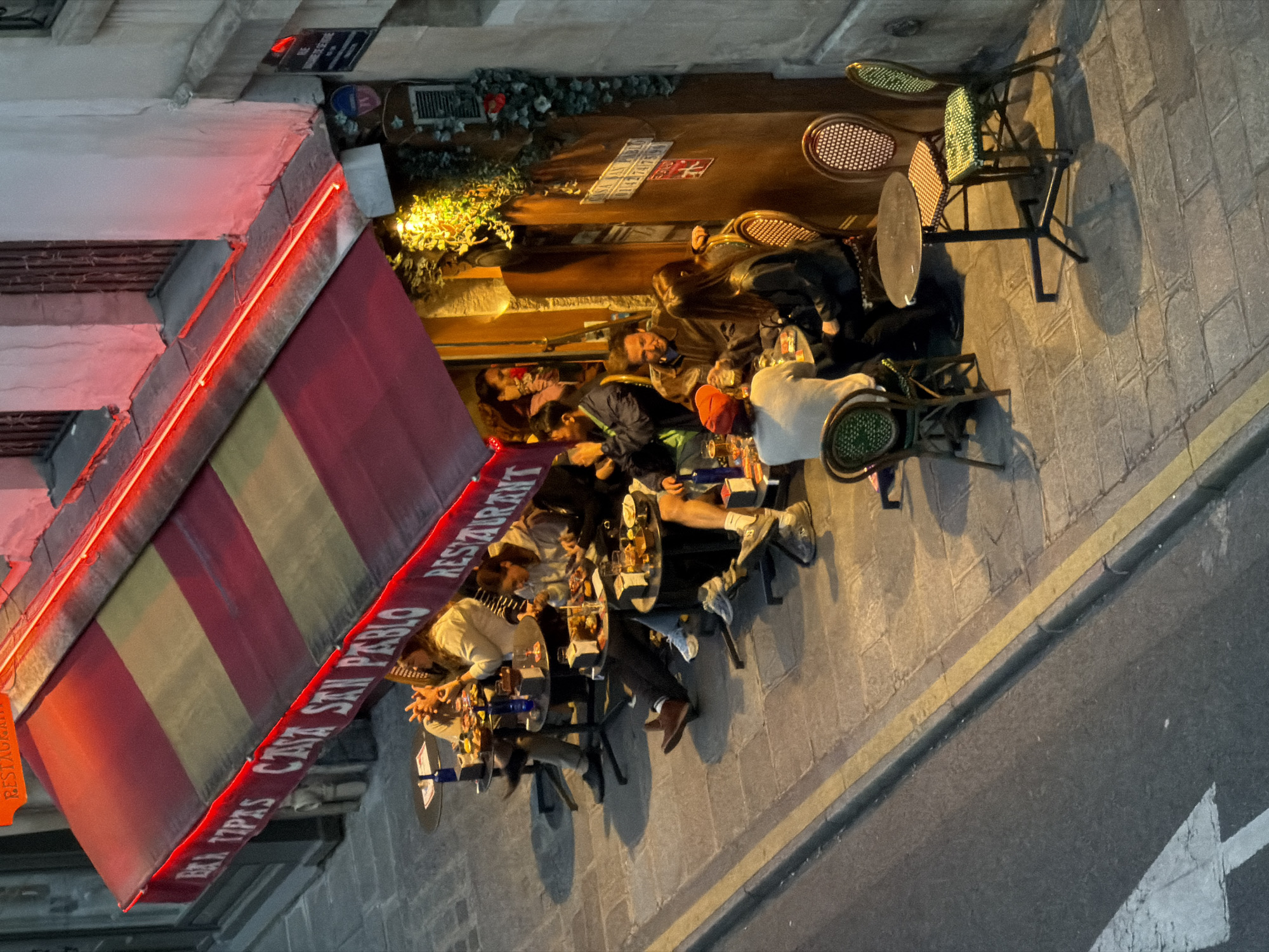 The image shows an outdoor dining area of the restaurant "Casa San Pablo" in Paris, France. The restaurant has a striped awning with the name of the restaurant written on it. Several people are seated at small, round tables along the sidewalk, appearing to be eating and socializing. The area is illuminated by ambient lighting from the restaurant and possibly streetlights. The sidewalk is paved, and a road runs parallel to the dining area with a faded white arrow painted on its surface. The restaurant's facade is adorned with plants and flowers above the door.