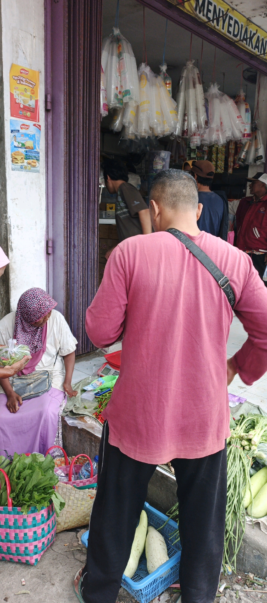 The image depicts a street scene in Kasiyan, Indonesia, focusing on a roadside market stall. In the foreground, a man wearing a pink t-shirt and black pants stands with his back to the camera, facing away from the viewer. To the left, a woman wearing a pink and white patterned hijab and a white shirt sits on a low wall, tending to produce. She is holding a plastic bag containing green vegetables. In front of her are two woven baskets, one containing leafy green vegetables and the other a plastic bottle. Behind the woman, a purple metal accordion gate is visible, with posters attached to the wall to its left.
The stall itself features an assortment of fresh produce laid out on a green tarp and a red scale. Bunches of long green beans and several light-colored gourds are prominent. Other items, possibly condiments or packaged goods, are visible on shelves behind the stall. The ground is paved and appears somewhat worn. The overall atmosphere suggests a busy, local marketplace.