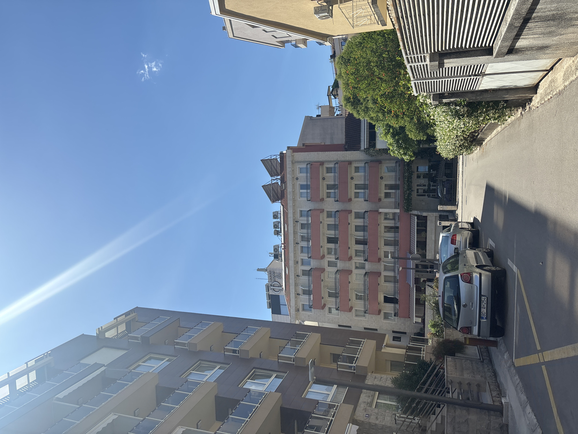 The image depicts a street scene in Budva, Montenegro. The angle is shot upwards from street level. The sky is clear and blue with a wisp of cloud visible. The buildings on either side of the street are multistory and of modern construction. Parked on the right side of the street are two silver cars. The license plate of the closer car reads "BD AH264". Foliage is visible interspersed between the buildings.