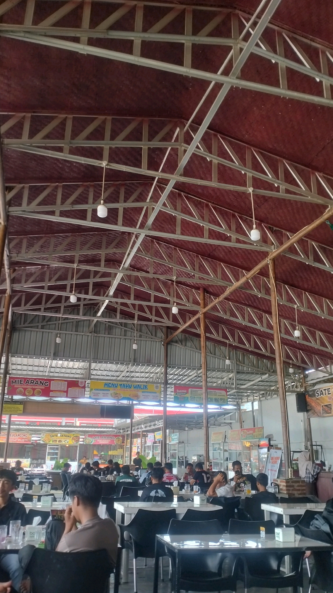 The image shows an outdoor food court in Nunangantara, Indonesia, with a large, open-air structure providing shade over numerous tables and chairs. The roof is supported by a metal framework and covered with red corrugated material. Several people are seated at tables, appearing to be dining or socializing. Signs advertising food items like "MIE ARANG," "MENU TAHU WALIK," and "SATE" are visible. The environment is casual and bustling, typical of a local eatery.