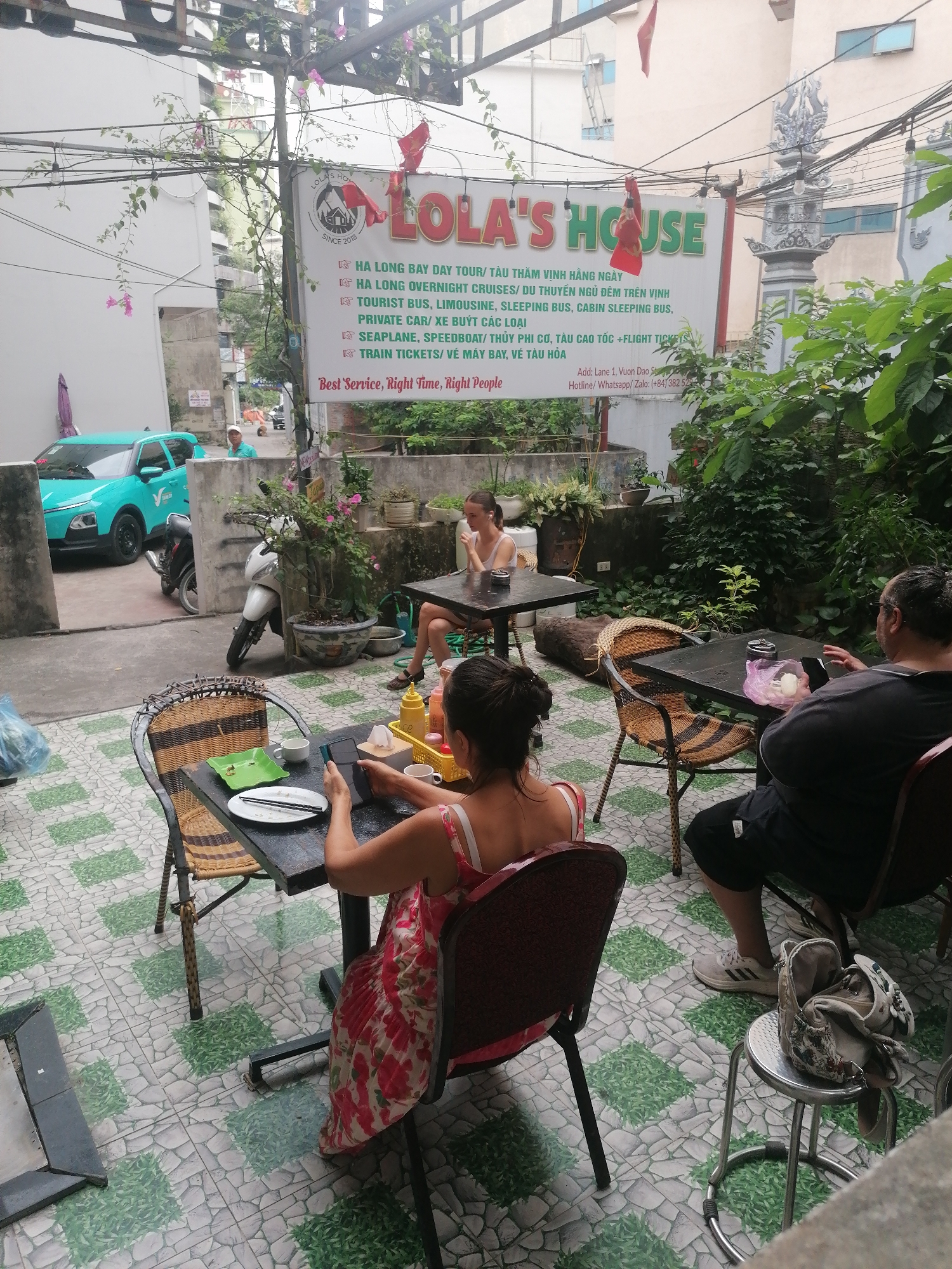 An outdoor seating area in Bai Chay Ward, Vietnam, hosts three individuals relaxing at tables during daytime. In the foreground, a woman with her back to the camera, wearing a red and floral dress, sits at a dark table with a white plate, chopsticks, and a phone in her hand. Beside her is a condiment caddy. To her right, a man in a dark t-shirt and shorts, also with his back to the camera, is engrossed in his phone. An ashtray and a small bag are on his table. Further back on the left, another woman in a white top sits at a table, holding a cup.
The scene is set on a distinctive tiled floor with green and grey patterns, beneath an overhanging structure adorned with lush green vines and pink flowers. Abundant foliage and potted plants surround the tables, creating a verdant atmosphere. In the background, an alleyway shows a teal-colored car, possibly a taxi service, and two motorbikes parked. A man in a light blue shirt is visible further down the alley.
A prominent sign for "LOLA'S HOUSE" dominates the upper right of the image, advertising various travel services including "HA LONG BAY DAY TOUR," "HA LONG OVERNIGHT CRUISES," "TOURIST BUS, LIMOUSINE, SLEEPING BUS," "PRIVATE CAR/ XE BUÝT CÁC LOẠI," "SEAPLANE, SPEEDBOAT/ THỦY PHI CƠ, TÀU CAO TỐC +FLIGHT TICKETS," and "TRAIN TICKETS/ VÉ MÁY BAY, VÉ TÀU HỎA." The sign also states "SINCE 2018" and "Best Service, Right Time, Right People," with an address "Add: Lane 1, Vuon Dao S" and hotline details. The overall ambiance is casual and relaxed, indicative of a guesthouse, cafe, or tour operator's office.