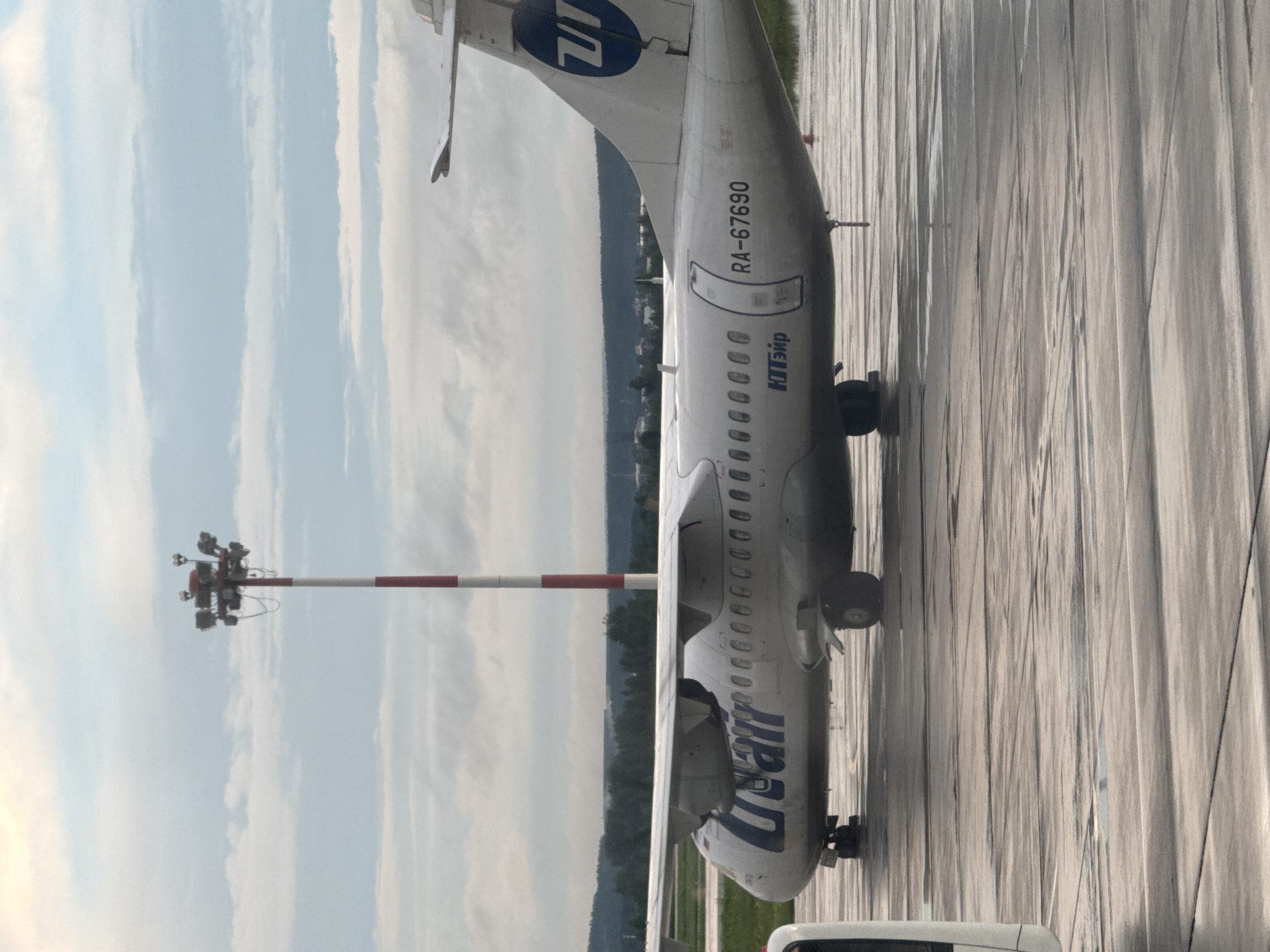 An aerial perspective shows a Utair ATR 72-500 aircraft, identified by the registration RA-67690, parked on a wet tarmac. The aircraft's white fuselage features the blue Utair logo on the tail and "Utair" lettering on the side. Red and white striped lights stand tall in the background, and a line of trees forms the horizon. The cloudy sky above contributes to the overall overcast atmosphere.