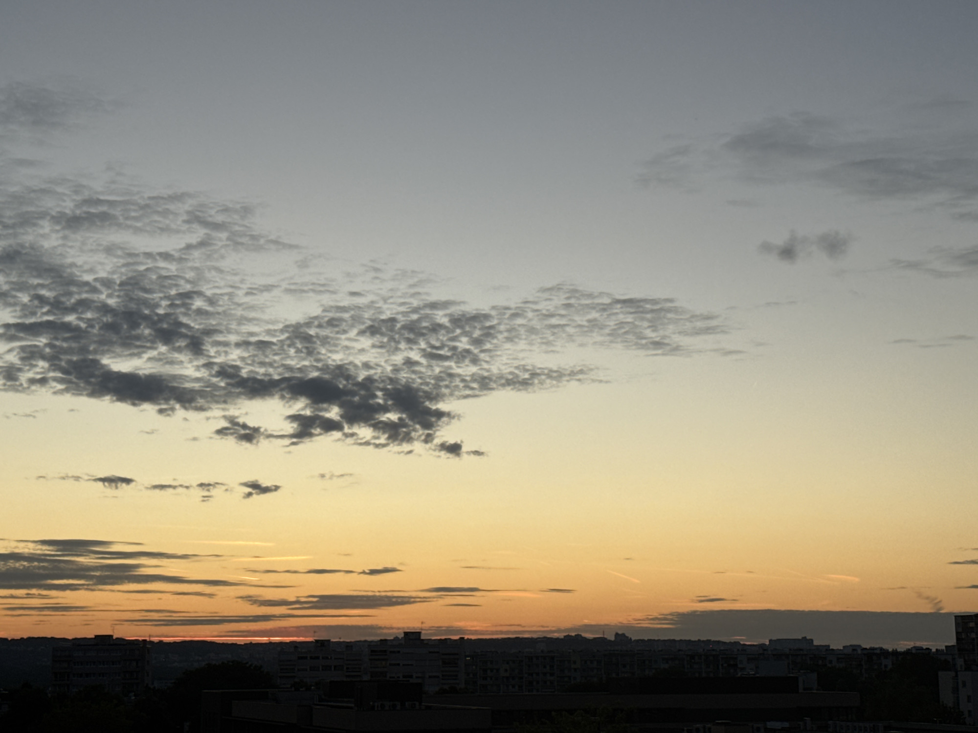 A low-angle, wide shot captures a cityscape at dawn or dusk, with a gradient sky transitioning from pale blue to warm orange. A band of dark, textured clouds stretches across the left side of the frame, with scattered wispy clouds and contrails visible higher up. Silhouetted buildings form a dark, uneven line along the horizon, with a few taller structures standing out. The overall impression is one of urban tranquility as day transitions to night or vice versa.
