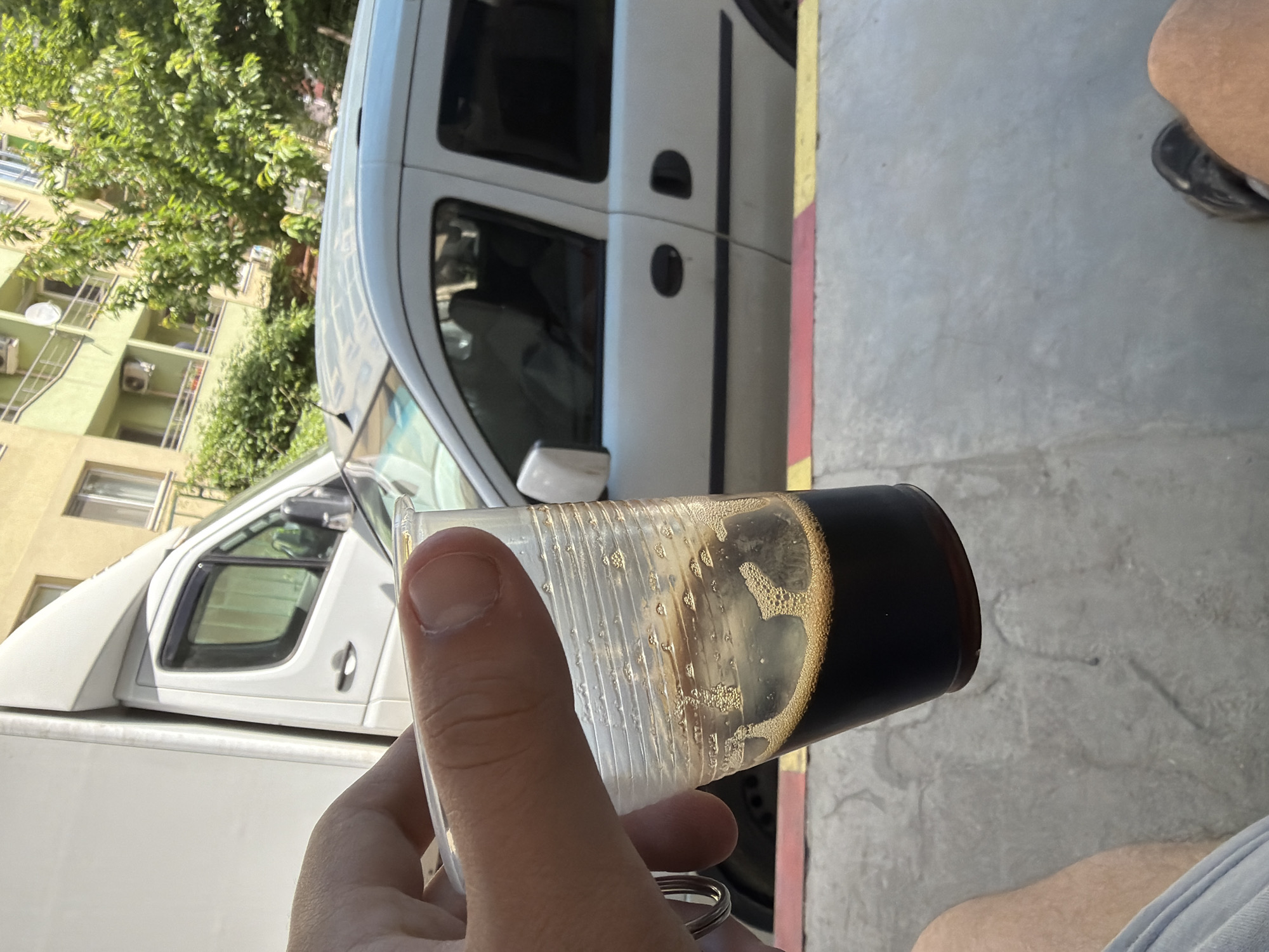 A person is holding a plastic glass filled with a dark liquid, possibly beer, with visible bubbles. The background shows a white van parked on a street with a building covered in greenery in the distance. The scene appears to be located in an urban area, possibly Sofia, Bulgaria. A small portion of a person's leg and shoe is visible to the right of the glass. The ground is concrete. The photo is taken from a first-person perspective.
