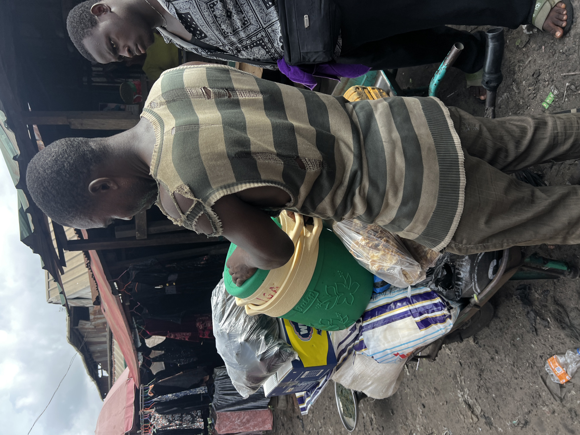 This media file captures a moment in an outdoor market in Nkpor, Nigeria, featuring two men and various goods. The image is oriented sideways, but depicts the subjects standing upright.
In the foreground, a man with short, dark hair is seen from the side, looking downwards. He wears a visibly worn, sleeveless vest with horizontal grey and tan stripes, showing several tears and frayed edges. He also wears dark, possibly striped, trousers. He is holding a distinctive green and yellow plastic container, possibly a bucket or cooler, with a floral pattern embossed on the green section and the word "Tulip" visible on the yellow rim. Strapped or carried with this container are several clear plastic bags, one of which contains what appears to be bread or pastries. Below the container, a yellow package with a prominent "M" logo is visible, alongside other bundled items and bags.
Behind him, another man is partially visible, seen from the chest up and looking towards the right side of the frame. He wears a black and white patterned shirt. His feet, clad in green and black Adidas brand sandals, are visible near the bottom right, suggesting he is standing near the foreground man.
The setting is a bustling outdoor market, characterized by a dirt ground and makeshift stalls or shops with corrugated metal roofs and fabric awnings in the background. The sky is overcast, indicating daytime. The scene conveys a sense of daily life and commerce in a busy urban environment. Litter, including a plastic bottle with partial text visible, is scattered on the ground.
