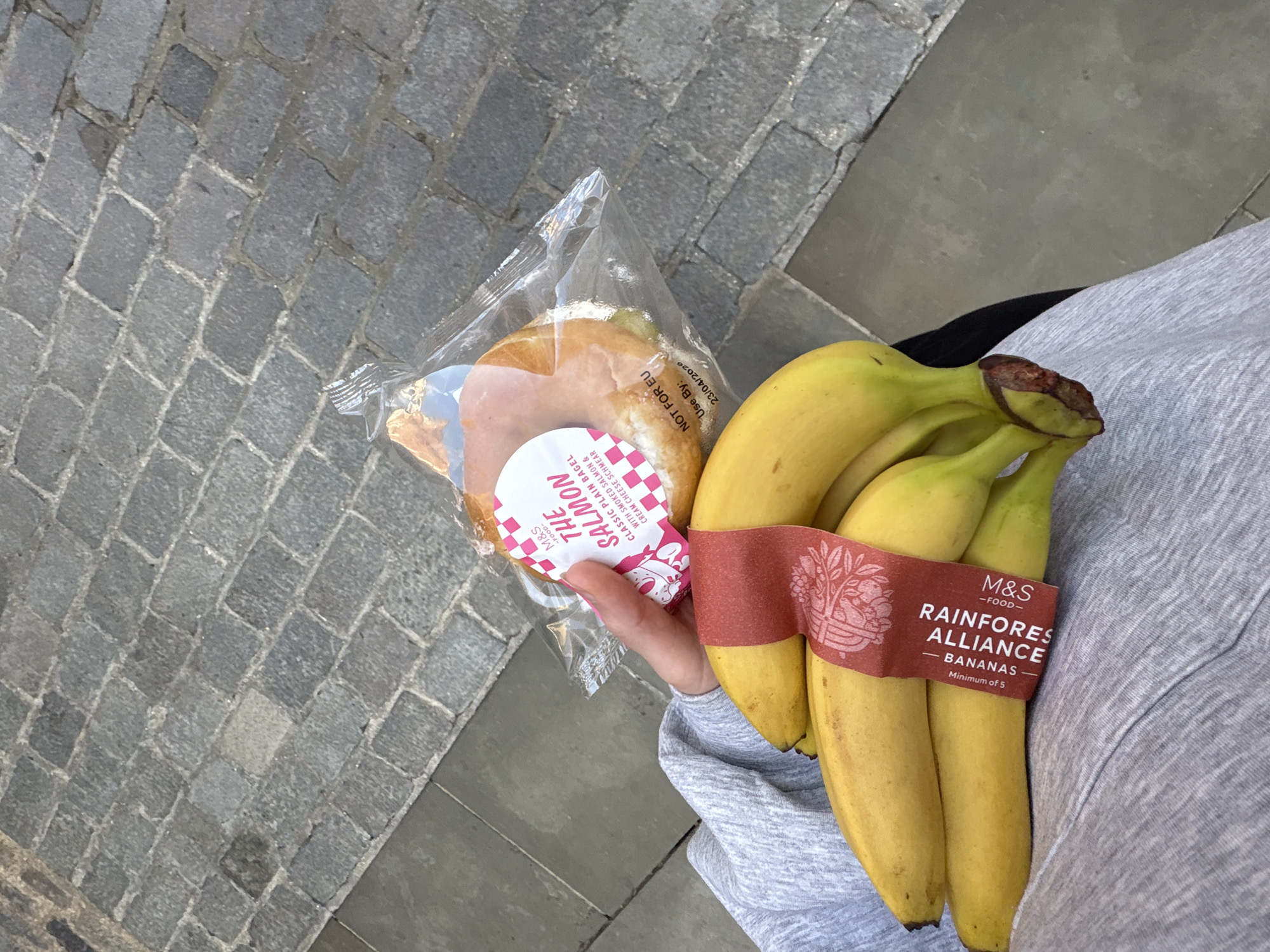 An overhead view captures a person holding a packaged meal and bananas while standing on a paved outdoor surface in London, United Kingdom. The person's right arm, clad in a light grey long-sleeved top, is visible, with their hand grasping two food items.
In their left hand, they hold a plastic-wrapped bagel sandwich. The visible label identifies it as "M&S -FOOD- THE SALMON CLASSIC PLAIN BAGEL WITH SMOKED SALMON & CREAM CHEESE SCHMEAR." Additional text on the packaging indicates "NOT FOR EU" and a "Use By: 23/04/2020" date.
Clutched in their right hand is a bunch of ripe yellow bananas, held together by a reddish-brown paper band. This band prominently displays "M&S -FOOD-" and states "RAINFOREST ALLIANCE BANANAS Minimum of 5." There appear to be around five to six bananas in the bunch.
The background shows a textured ground made of dark grey cobblestone or paving stones, suggesting an urban street or pathway. The scene is illuminated by daylight, and the ground appears dry. No specific emotions are discernible, but the image conveys a moment of carrying purchased food items.