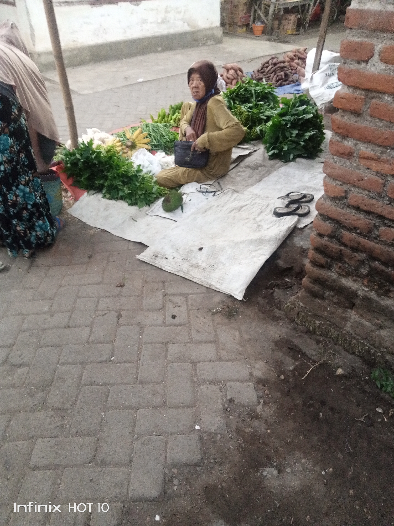 The image depicts an outdoor market scene in Sono Ageng, Indonesia. In the foreground, a woman wearing a brown headscarf and a tan long-sleeved shirt is sitting on a white tarp. She is holding a dark-colored bag in her lap. Next to her are piles of fresh green vegetables, including what appear to be bunches of leafy greens and some cucumbers. In the background, a large pile of what look like sweet potatoes or yams is visible, along with some cardboard boxes and what might be a wooden stall. The ground is paved with gray interlocking bricks. To the right of the scene, a brick wall is partially visible, showing reddish-brown bricks. A pair of black flip-flops rests on the tarp near the woman. The overall lighting suggests it is daytime.