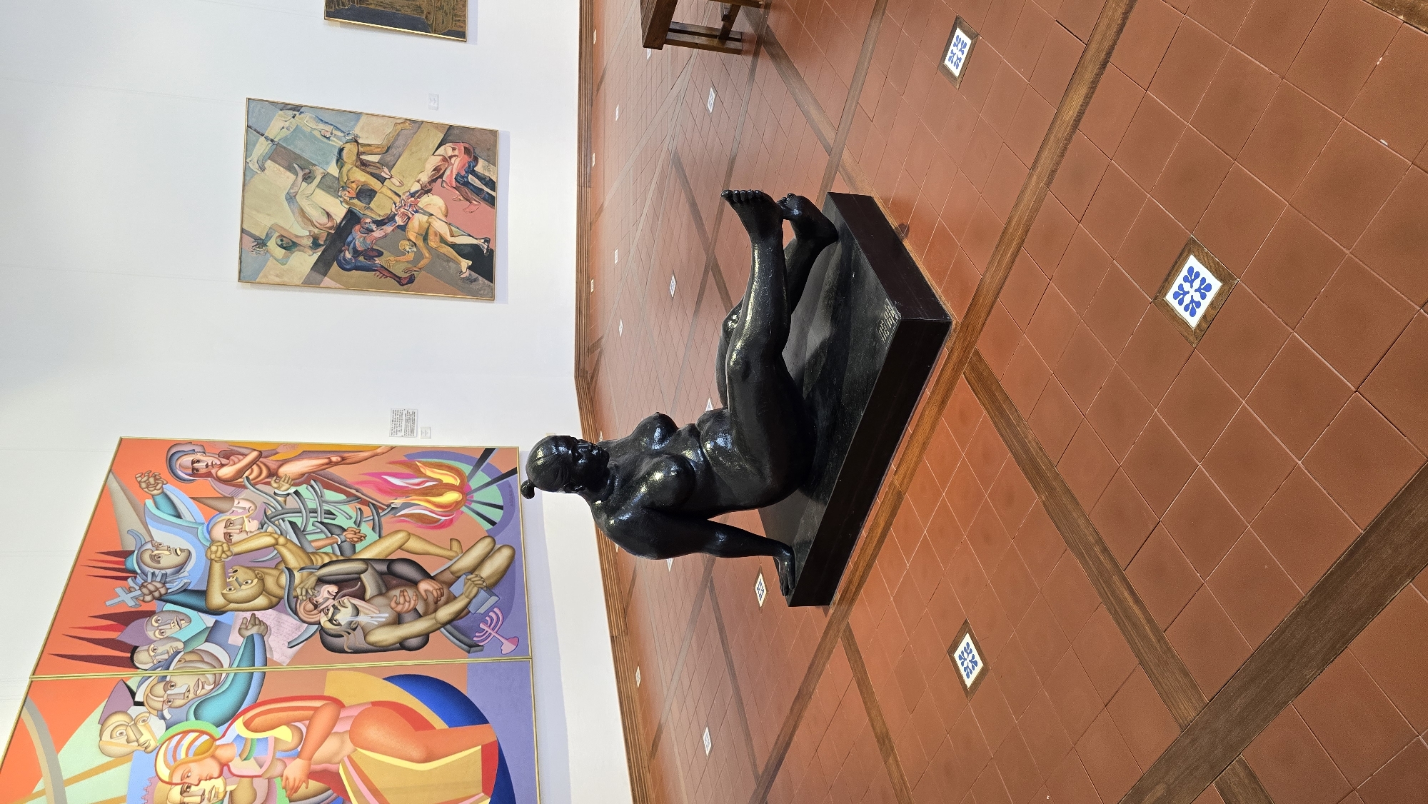 The image displays a museum exhibition hall with terracotta tiled flooring and white walls. In the foreground, a dark, possibly bronze, sculpture of a nude woman is seated on a black rectangular plinth. The sculpture depicts a full-figured woman with her legs extended and slightly bent, her arms resting on the ground. Her head is turned to her right.
In the background, two large paintings are hung on the wall. The painting on the left is a diptych with vibrant colors, depicting numerous figures in a dynamic and possibly chaotic scene with flames. The painting on the right is smaller and displays several reclining nude figures in muted colors, arranged in a somewhat abstract geometric pattern.
The environment suggests an indoor art gallery setting. The flooring features a pattern of terracotta tiles with inlaid wooden strips forming diagonal lines and a central decorative tile pattern of blue and white squares. Part of a wooden bench is visible on the far right edge of the image. There are no people visibly interacting with the artworks or the sculpture in the frame. The location is identified as the Hof HaCarmel Regional Council, Israel, implying this is likely a museum or cultural center within that region.