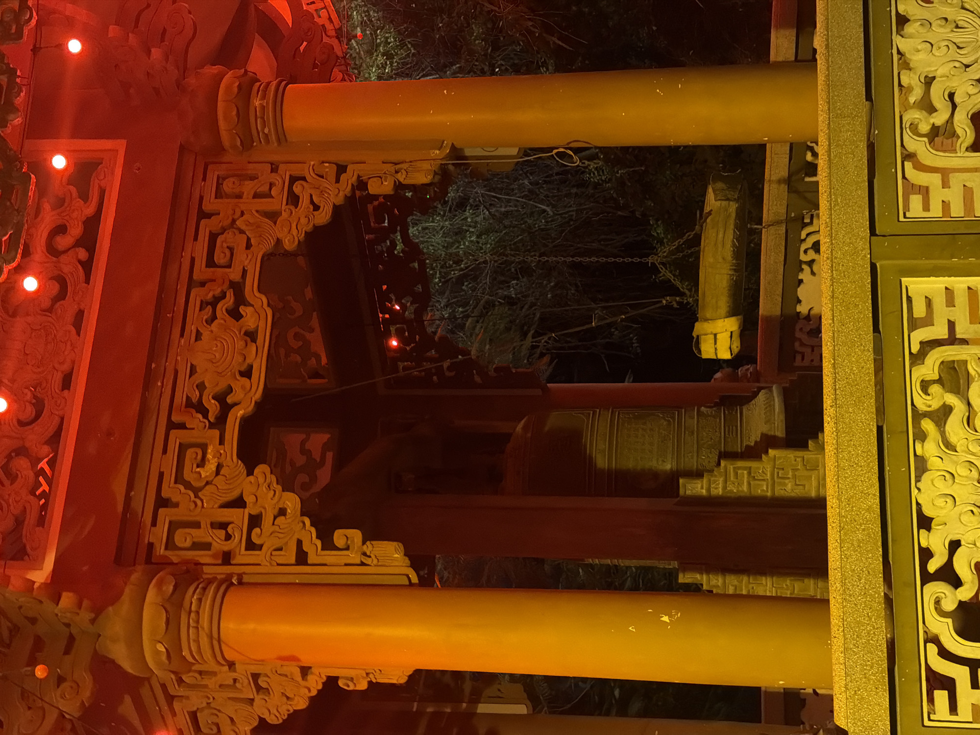 The image shows a temple structure at night in Nha Trang, Vietnam, illuminated by red lights. Large, ornate wooden columns and decorative eaves are visible. A large bell, likely a temple bell, hangs within the structure, along with a wooden beam suspended by chains. The background consists of dark foliage, suggesting an outdoor setting within a garden or temple grounds. The foreground features a yellow, patterned railing. There are no people or animals present in the image.