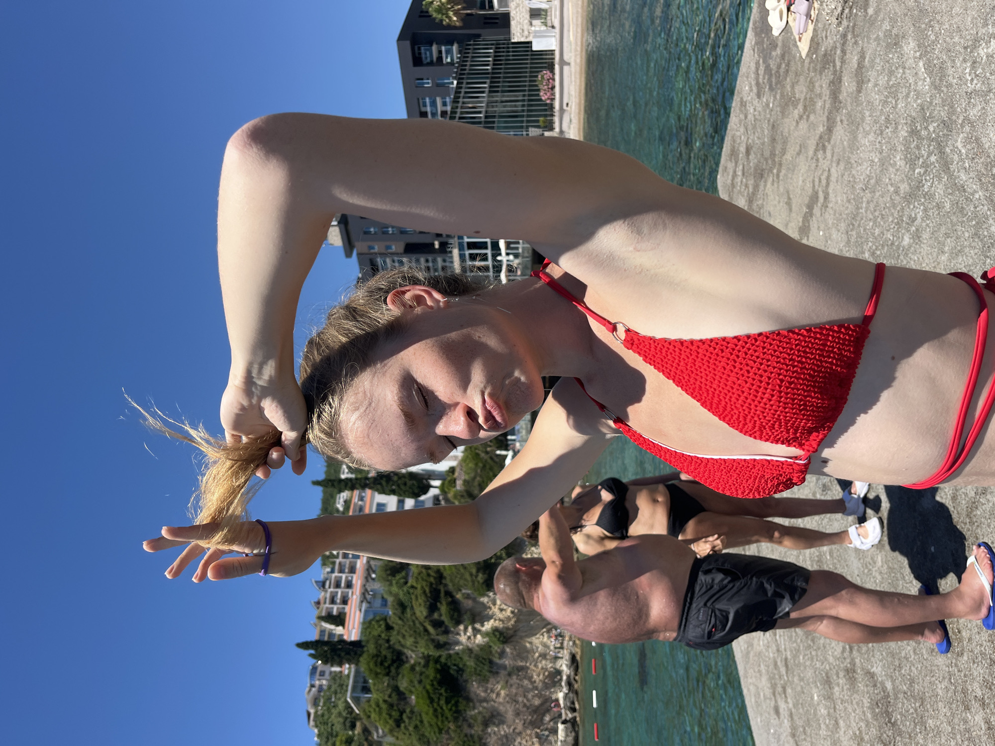 This image depicts a sunny day on a pier in Budva, Montenegro. In the foreground, a young woman in a red bikini is posing with her hand in her blonde hair, her lips puckered. Behind her, a man in black swim trunks and a woman in a black bikini stand facing away from the camera. The clear blue water of the Adriatic Sea is visible, with buildings and lush greenery clinging to the hillside in the background. The overall scene is one of leisure and enjoyment on a summer day by the coast.