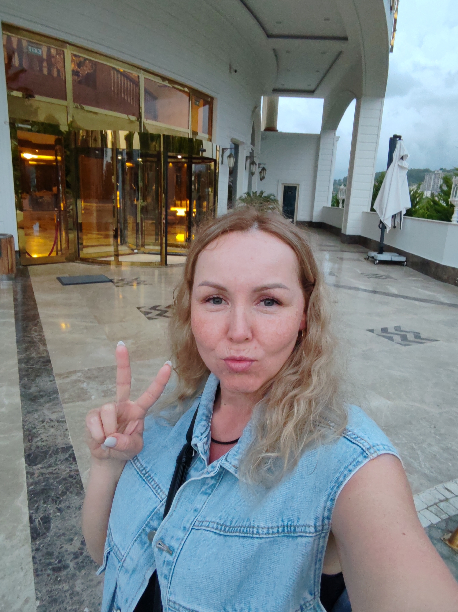 The photograph captures a light-skinned woman with blond, wavy hair who appears to be in her late 30s to mid 40s. She is wearing a sleeveless denim jacket over a black garment. Her left hand is raised, forming a peace sign, while she purses her lips into a pout.
The setting is outdoors, possibly at the entrance or terrace of an upscale establishment. Behind her is a revolving door entrance with golden trim and reflective glass, suggesting a hotel or resort. The area is paved with marble flooring with decorative patterns. To her right, there's a covered walkway or veranda with white pillars and a partially visible view of trees and buildings. A white patio umbrella on a stand is also visible.
While I lack the specific data to pinpoint the exact location, given the architectural style and landscape, it is highly probable that this photograph was taken in Alanya, Turkey.