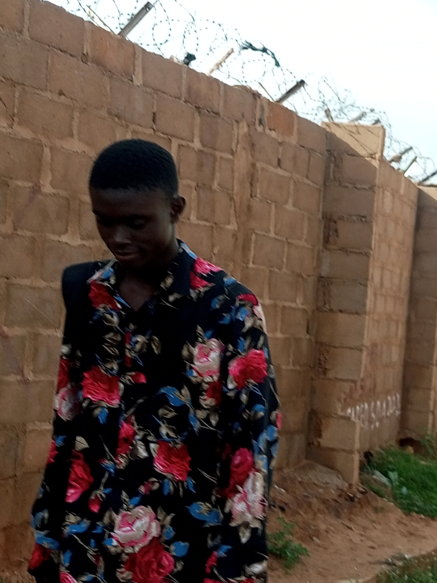 A young Black male with short hair stands outdoors in front of a brick wall topped with barbed wire. He is wearing a black button-down shirt with a large floral pattern of red, pink, white, and blue flowers and green leaves. The brick wall appears to be made of light brown, rectangular blocks, and there is some dry grass and dirt on the ground in front of it. The sky is visible above the wall, and it appears to be overcast. The scene suggests an outdoor location within the city of Gombe, Nigeria.