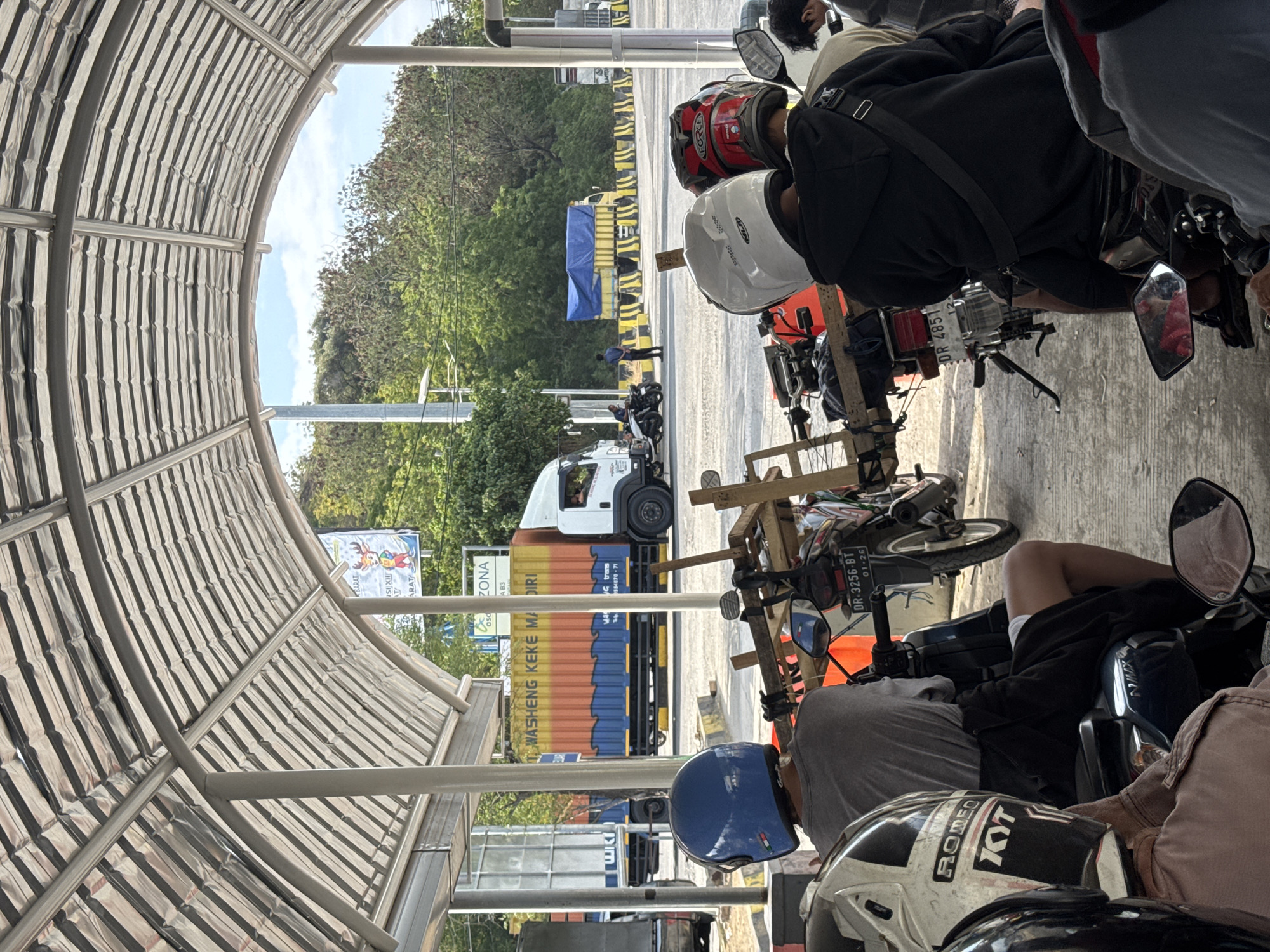 The image depicts a queue of motorcycles and a truck at a location resembling a port or ferry terminal in Upt Tambak Sari, Indonesia. A covered structure with a corrugated metal roof and metal beams extends over the scene, suggesting it might be a waiting area or loading bay.
In the foreground, three individuals are seated on motorcycles, viewed from behind. The person on the left wears a blue helmet and a grey t-shirt. The person in the middle wears a white helmet and a black hoodie with a crossbody bag. The person on the right wears a red and black helmet. Their motorcycles are parked in a line, some with wooden structures attached.
In the background, a large white and orange truck with the text "WASHENG KEKE MANDIRI" is visible, along with other vehicles and trees. The environment appears to be outdoors, with a bright sky and sunlight casting shadows. The ground is paved. A sign with the word "ZONA" and a cartoon deer can be seen on a pole. The presence of multiple vehicles, especially motorcycles, suggests this is a point of transit.