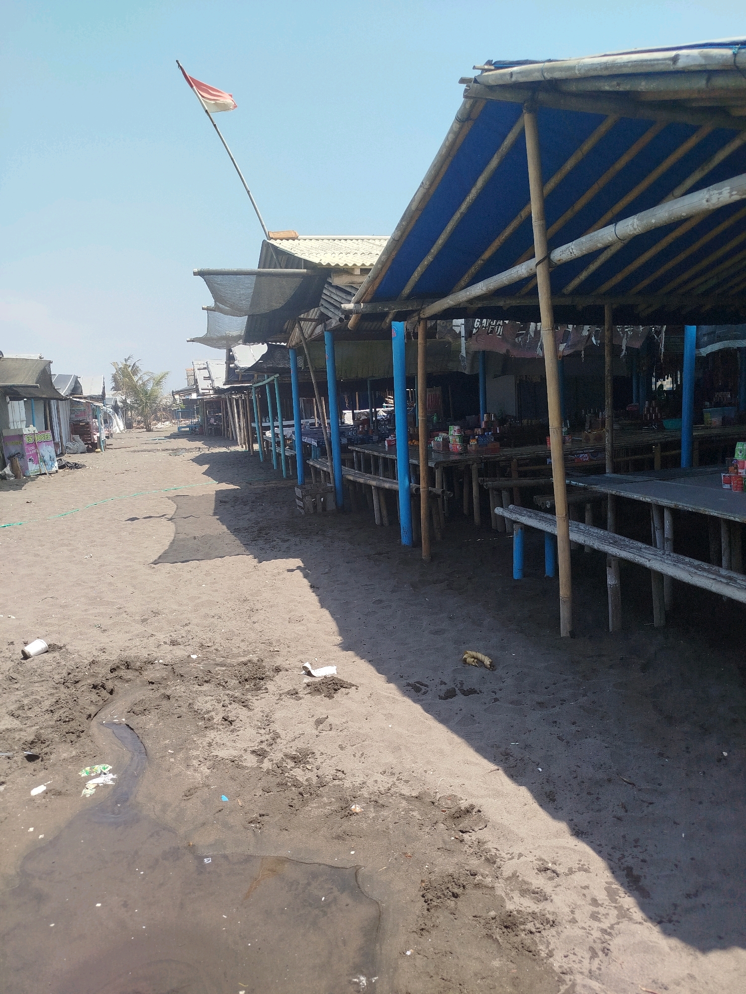 The image depicts a row of simple, open-air food stalls constructed with bamboo and covered by blue tarps on a sandy beach. The stalls are lined up along a sandy pathway. In the foreground, the sand is wet in some areas, with a small stream of water meandering through it and some scattered litter. In the background, more stalls and structures are visible, extending into the distance under a clear blue sky. A red and white flag, likely the Indonesian flag, is flying from a tall pole. The overall scene appears to be a beachside market or dining area.