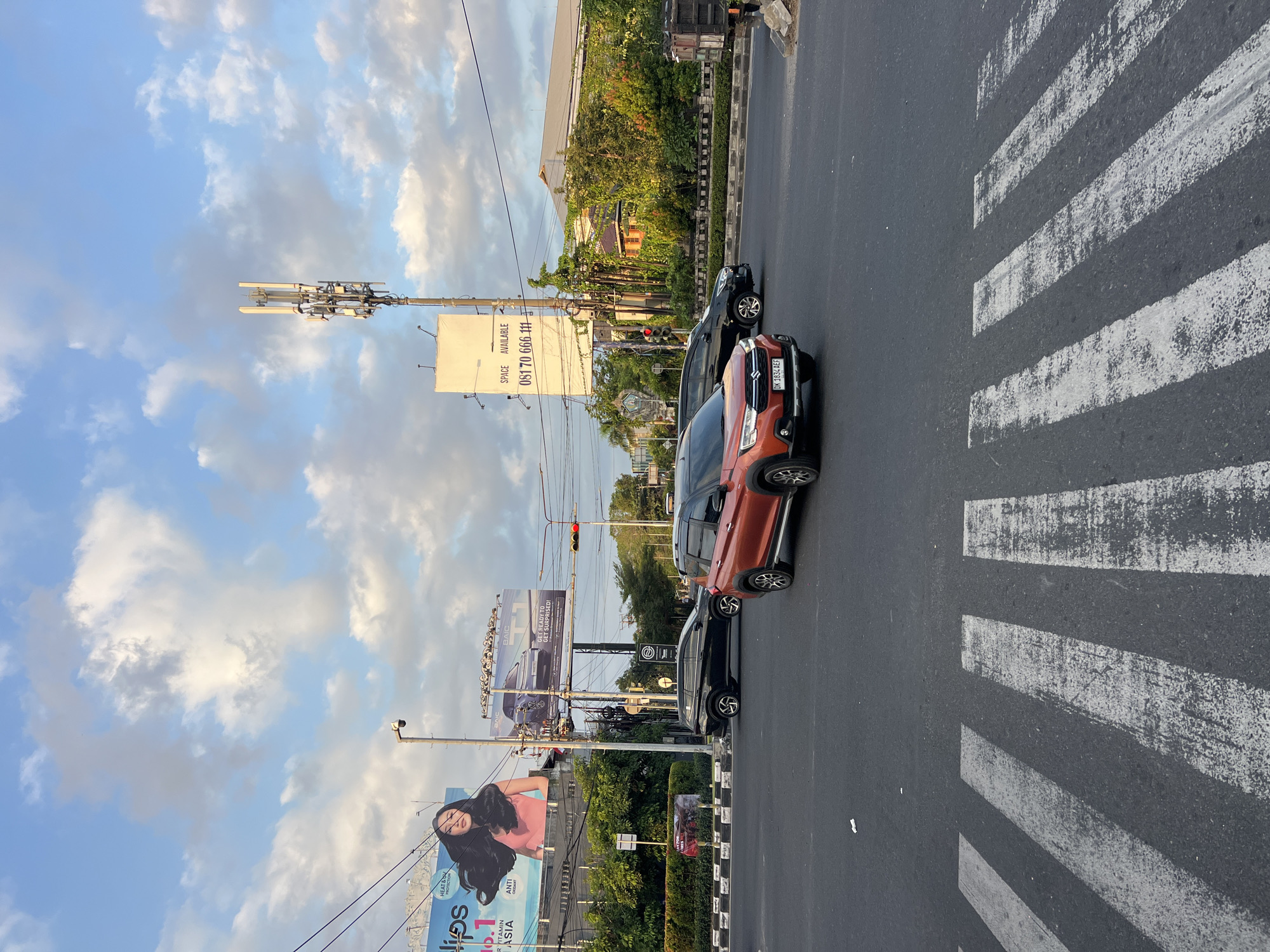 An over-the-shoulder shot captures a road intersection in Denpasar Barat, Indonesia, under a partly cloudy sky during the late afternoon. The foreground is dominated by a pedestrian crossing with bold white stripes painted on the asphalt. Two vehicles are visible: a striking orange SUV in the middle lane, facing left, and a black sedan slightly behind it, also facing left. The orange SUV has a license plate reading "DK 1834 AEF". In the background, a large billboard advertises "LIPS" with an image of a woman and the words "HEAT & DRY PROTECTION" and "ANTI AGING", alongside another billboard for BAIC vehicles, promoting the "T11" model with the tagline "GET READY TO GET SURPRISED!". A cell tower stands tall behind the billboards, and traffic lights are visible, with one light showing red. Lush green trees and buildings line the street in the distance, contributing to the urban landscape. The scene suggests a moment of everyday urban life in Denpasar Barat, with traffic flowing through the intersection.