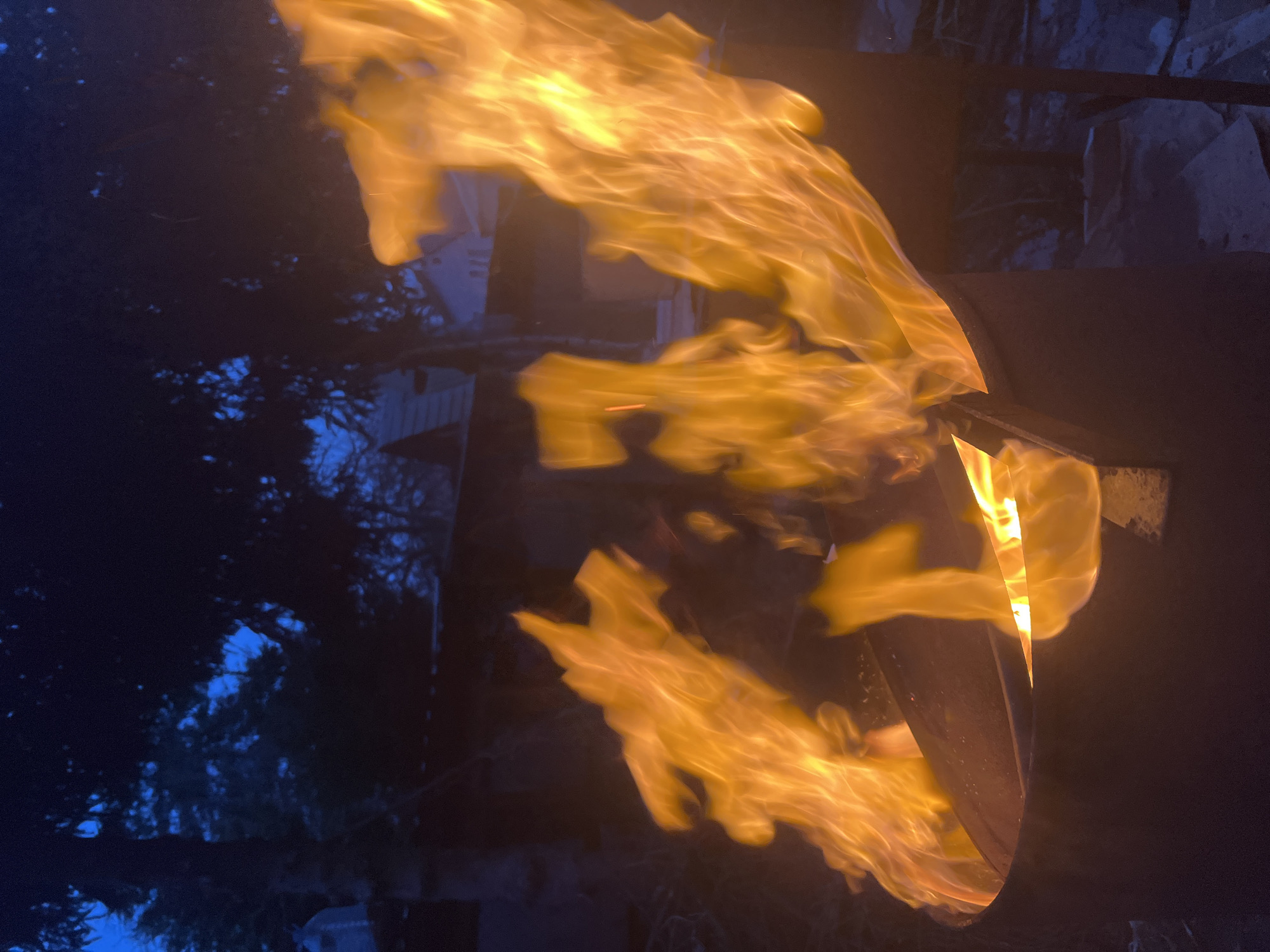 The image displays a vibrant, active fire burning in a dark outdoor setting, likely at night or dusk. Large, bright orange and yellow flames dominate the right side and center of the frame, swirling and extending upwards. These dynamic flames appear to be emanating from a dark, metallic object, possibly a fire pit or barrel, partially visible on the right.
The background is predominantly dark blue and black, indicating the time of day. Silhouetted against this dark sky are numerous trees and dense foliage, suggesting a natural or semi-natural environment such as a backyard or wooded area. Hints of a light-colored structure, possibly a building or fence, are faintly visible behind the flames and trees on the left side, reflecting some light. The image strongly emphasizes the intense glow and movement of the fire against the dark surroundings. No people or visible text are present in the scene.