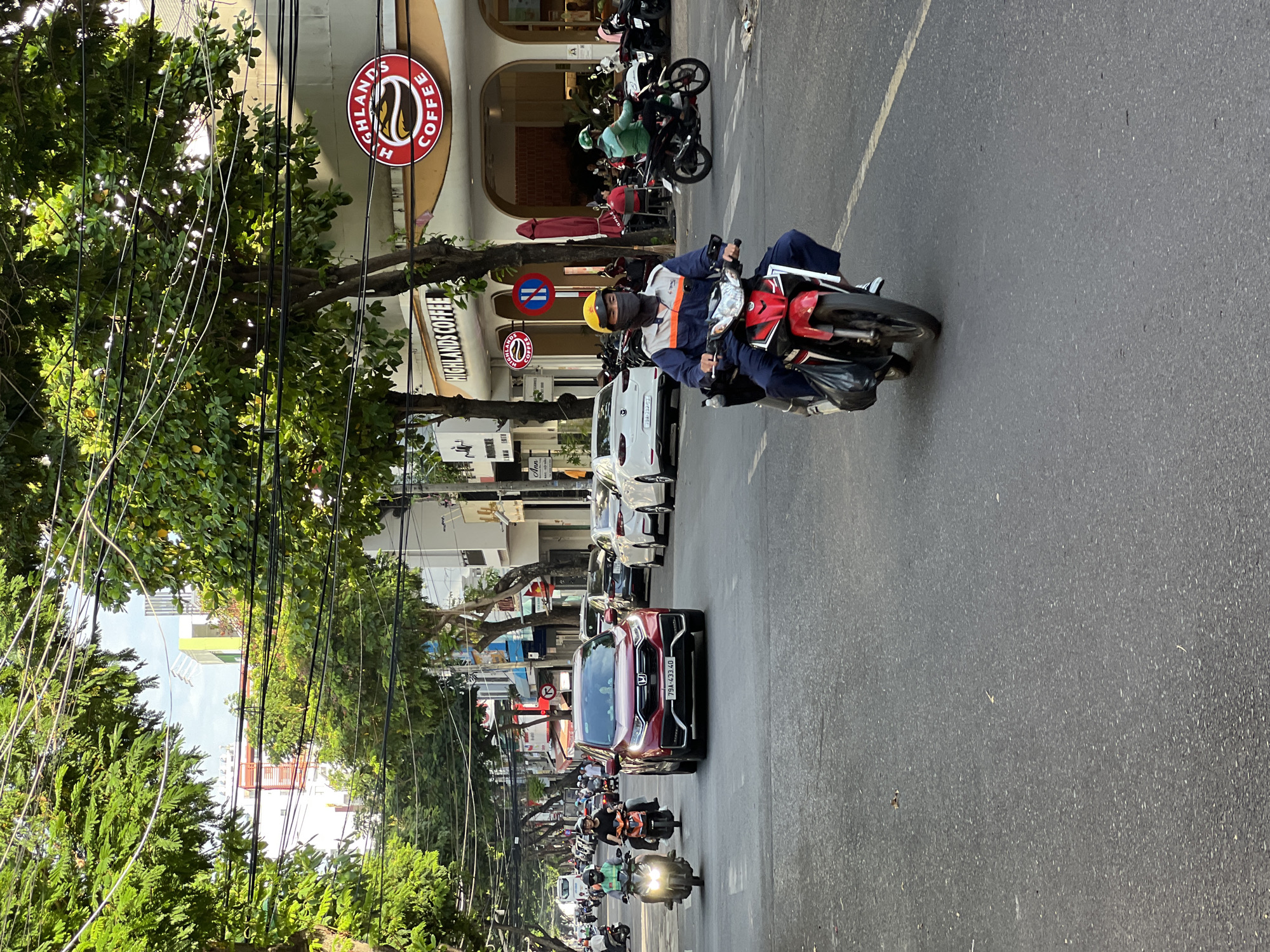 A busy street scene in Nha Trang, Vietnam, is depicted, with a focus on traffic and commercial establishments. The foreground features a man on a red and white motorcycle, wearing a blue and orange uniform, a yellow helmet, and a black mask, riding down the asphalt road. The road is marked with a yellow line. To the left, a red SUV is stopped, and further back, several motorcycles are in motion. On the right side of the street, a "Highlands Coffee" establishment is visible, with its distinctive logo. Buildings line the street, with trees and overhead wires creating a canopy. The lighting suggests it is daytime, possibly late afternoon, with shadows cast on the road. The overall impression is one of urban activity and commerce.