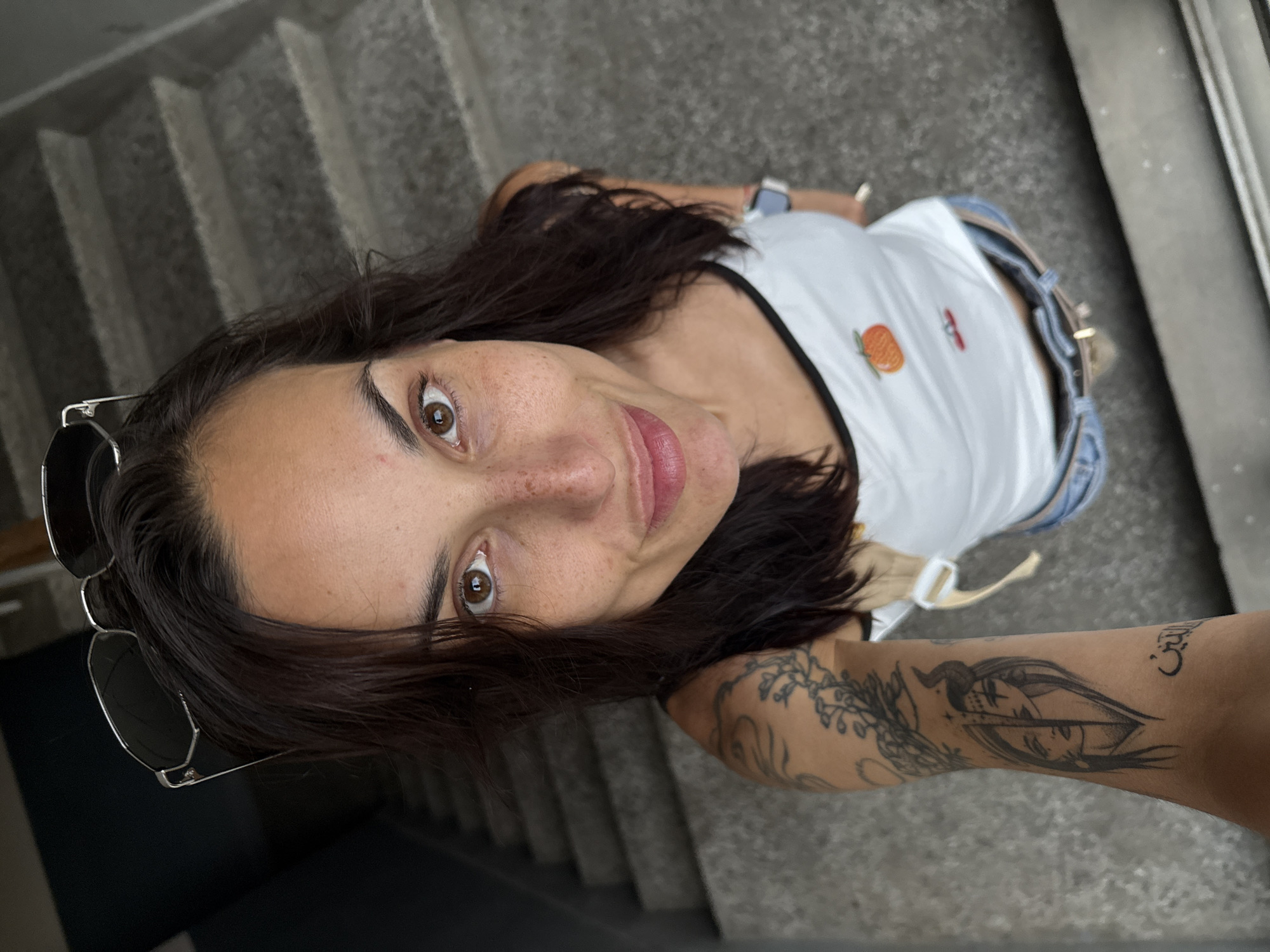A female adult is depicted in a high-angle selfie, looking directly into the camera with a slight smile. She has dark brown, wavy hair, brown eyes, and prominent freckles across her nose and cheeks. Silver-framed sunglasses are pushed up onto her head. She wears a white tank top with black trim and a small orange fruit emblem on the chest, light-wash denim shorts, and a light-colored belt with a metal buckle. A watch is visible on her left wrist. Her left arm displays multiple tattoos: a floral/vine design on the upper arm and a split-face tattoo on the forearm depicting one half as human/feminine and the other as horned/demonic with forehead symbols, accompanied by Arabic script below it. The background features a concrete or stone staircase ascending, indicating an indoor or sheltered outdoor stairwell environment. The scene is lit by natural light. This image is located in the city of Pleven, Bulgaria.