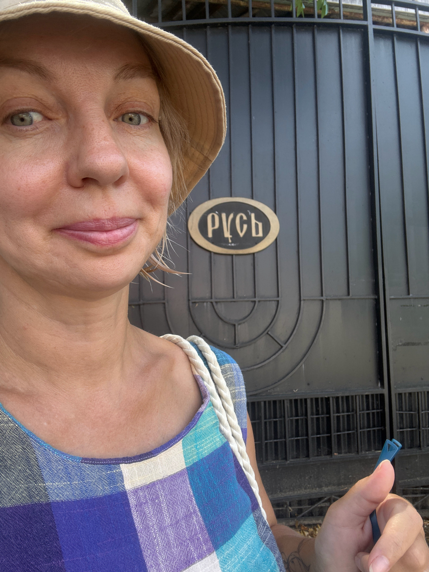 A woman wearing a straw hat and a blue and purple plaid dress takes a selfie in front of a black metal gate. On the gate, an oval-shaped plaque with Cyrillic script is visible. The plaque reads "РУСЬ". The woman is holding a small blue object in her right hand. The background appears to be outdoors, with foliage visible above the gate. The image is likely taken in Sochi, Russia, given the context of the Cyrillic script and the commonality of such gates in Russian cities.