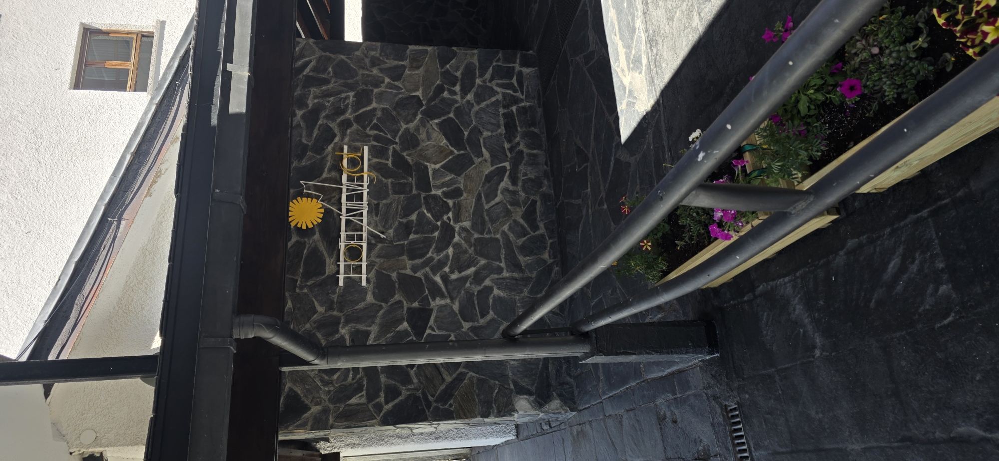 The image depicts the exterior of a building with a stone facade, likely a hotel, situated in Sallent de Gállego, Spain. A sign with white lettering and a yellow sun emblem is affixed to the stone wall, reading "HOTEL NIEVE". A dark metal railing runs diagonally across the foreground, bordering a planter box filled with various flowering plants, including pink petunias and yellow flowers. The ground is paved with dark stone tiles. A downspout is visible on the left side of the image, attached to a dark overhang. The building's architecture suggests a mountain or rustic setting, consistent with the Pyrenees region where Sallent de Gállego is located. The lighting indicates it is daytime, with shadows cast on the ground, suggesting the sun is at an angle. No people are present in the image.