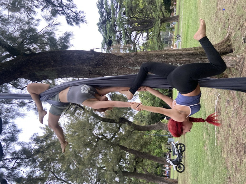 Two women engage in aerial acrobatics using a gray silk suspended from a tree. One woman wears a gray top and shorts, her legs intertwined in the silk. The other woman, with red braided hair, wears a blue crop top and black leggings, inverted while gripping the first woman's hands. A park setting with trees, grass, and people in the background, along with a parked scooter.