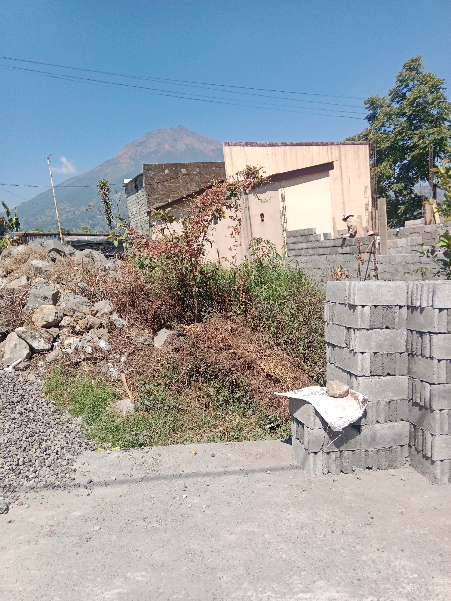 The image depicts an outdoor scene with a prominent volcano in the background. In the foreground, there is a construction site featuring stacks of grey concrete blocks. A person wearing a hat and light-colored clothing is partially visible, appearing to be working on the construction. To the left, there is a pile of large stones and debris, along with dry brush and some green grass. In the middle ground, there are several buildings, one of which has a light-colored facade. A tree with green foliage is visible on the right side of the image. The sky is clear and blue. The location is identified as Jurangjeru, Indonesia.