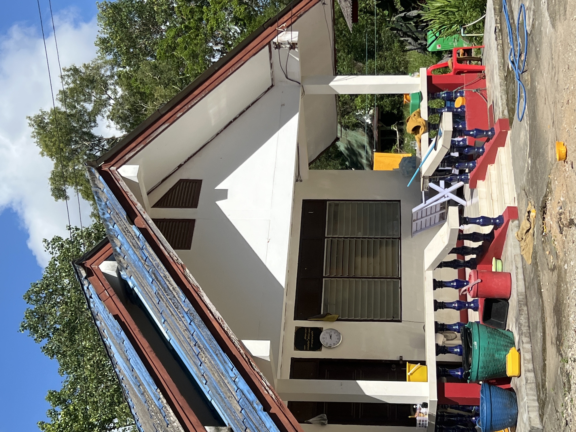 The image shows a light-colored, single-story house with a red and blue trim on the porch railing. The roof is made of blue corrugated metal with brown trim. A white column supports the porch overhang. There are several blue, green, and red buckets near the porch, as well as a yellow watering can and a black dustpan. A white folding chair is visible on the porch. The house is surrounded by lush green trees and vegetation. In the foreground, a blue hose lies coiled on the ground. The sky is blue with scattered white clouds. The scene appears to be located in a residential area in Thep Krasatti, Thailand, given the architectural style and vegetation. There are no people visible in the image.