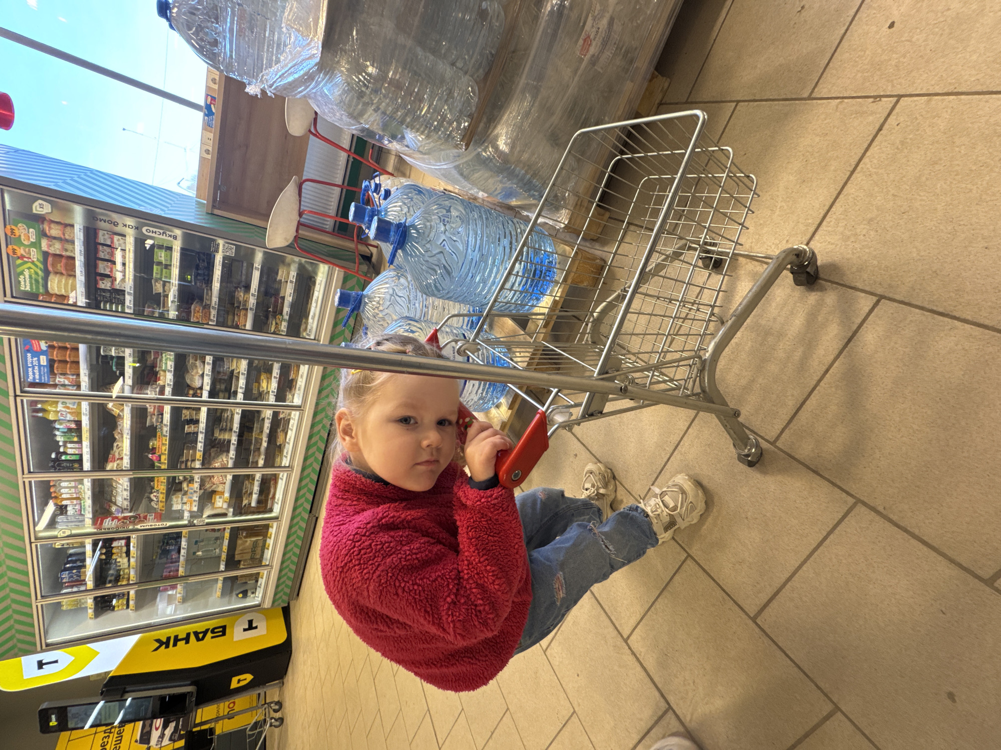 A young girl, wearing a red fuzzy jacket and blue jeans, is sitting in a wire shopping cart filled with large bottles of water. She is holding a red object in her hands. The shopping cart is on a tiled floor inside a store. Refrigerated display cases with food items are visible in the background. Signs in Russian, including one advertising "T Bank," indicate that the location is likely in Russia. The text also mentions "Perm" on one of the signs, indicating the city of Perm.