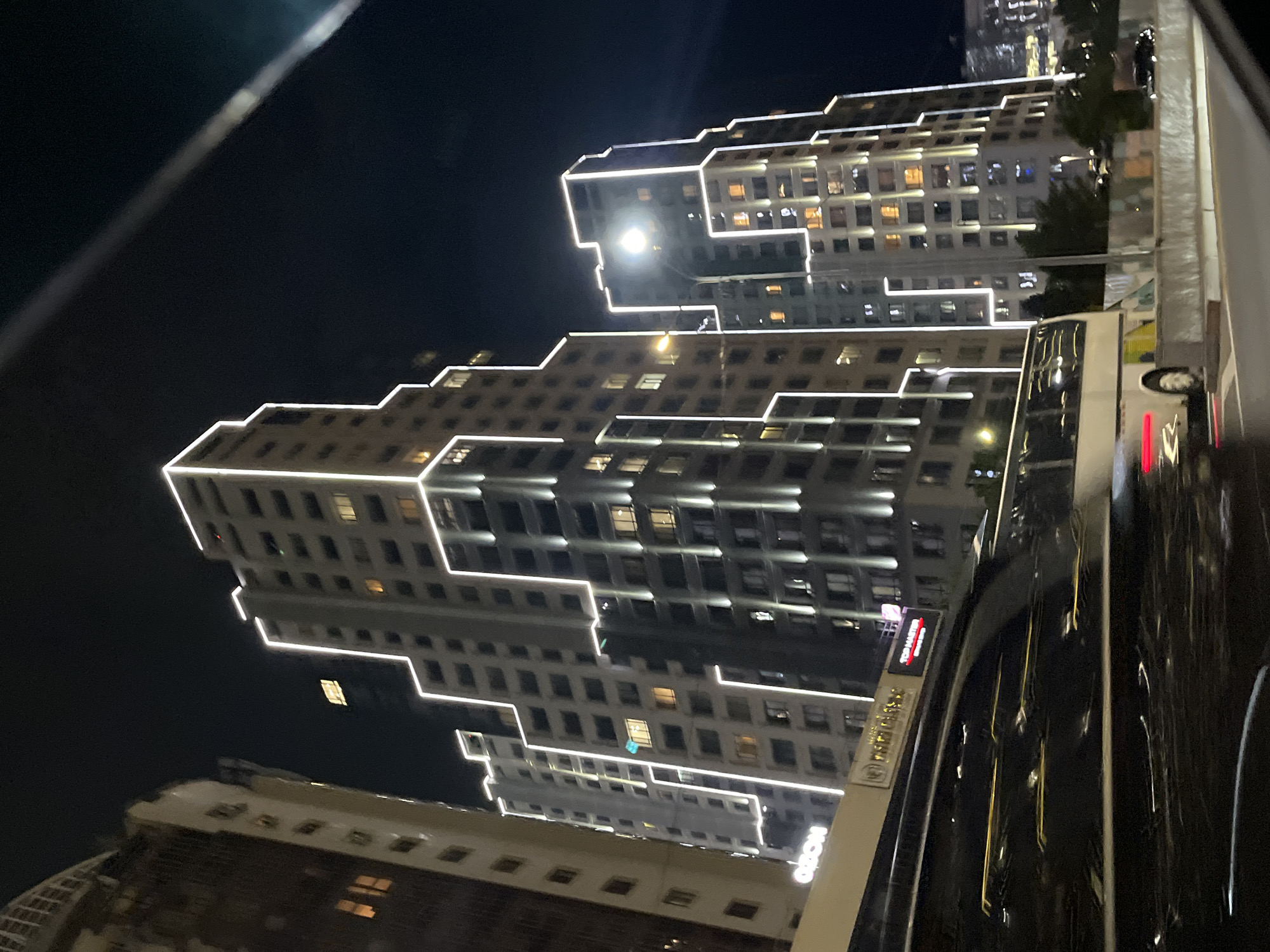 A night time, low-angle, eye-level, medium shot captures a modern urban landscape in Almaty, Kazakhstan, dominated by strikingly illuminated skyscrapers. The buildings, some of which appear to be residential complexes or office buildings, feature geometric architectural designs accented with bright white LED strip lighting that outlines their edges and patterns. Several windows in the buildings are lit with warm, orange light, suggesting occupancy.
In the foreground, a bus with a dark, reflective exterior is moving, its surface mirroring the lights and buildings around it, creating streaks of light due to motion blur. The bus is white with some colorful graphics visible on its side. It is traveling on a road bordered by a concrete barrier. Behind the bus and further into the mid-ground, other vehicles are visible, including cars, further contributing to the sense of an active urban environment.
The sky is a deep, dark blue, almost black, indicating it is nighttime. The overall impression is one of a vibrant and modern city at night, with the architectural lighting creating a dramatic visual effect. The scene appears to be taken from another vehicle, possibly a car, given the slight motion blur and the angle. The text "OZON" is visible on the side of the bus.