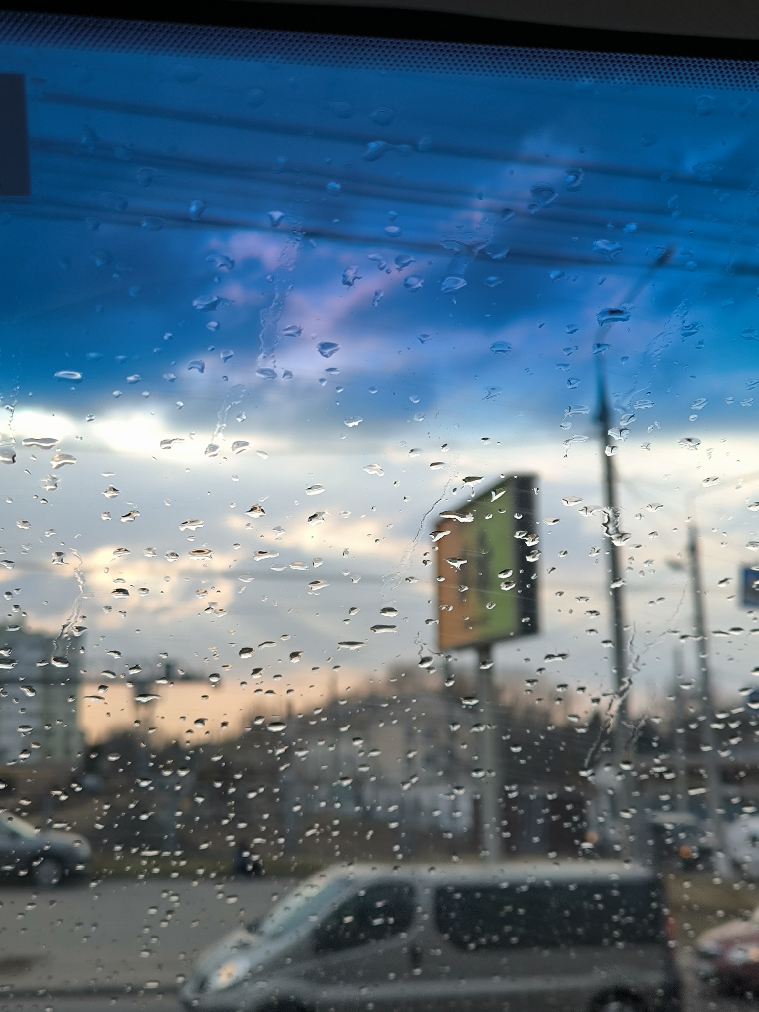 The image displays an urban street scene in Vinnytsia, Ukraine, observed from inside a vehicle through a rain-splattered windshield. The foreground is dominated by numerous water droplets of varying sizes adhering to the glass surface. The upper edge reveals a dark, textured vehicle interior component above a blue-tinted section of the windshield. The background elements are out of focus due to the narrow depth of field.
The sky in the upper background transitions from deep blue on the left to lighter shades of white and grey, with subtle hints of pink or orange, suggesting either dawn or dusk. Below the sky, indistinct multi-story buildings are visible. A prominent, rectangular advertising sign, presenting a green and orange/brown coloration, is situated in the mid-right. Multiple slender, vertical poles, consistent with streetlights or utility infrastructure, are scattered throughout the mid-right background. On what appears to be a road or street, several vehicles are present in blurred form, including a light-colored van or minibus centrally located in the lower midground, with other vehicle outlines to its left and right. No identifiable human figures are present. The scene indicates current or recent precipitation.